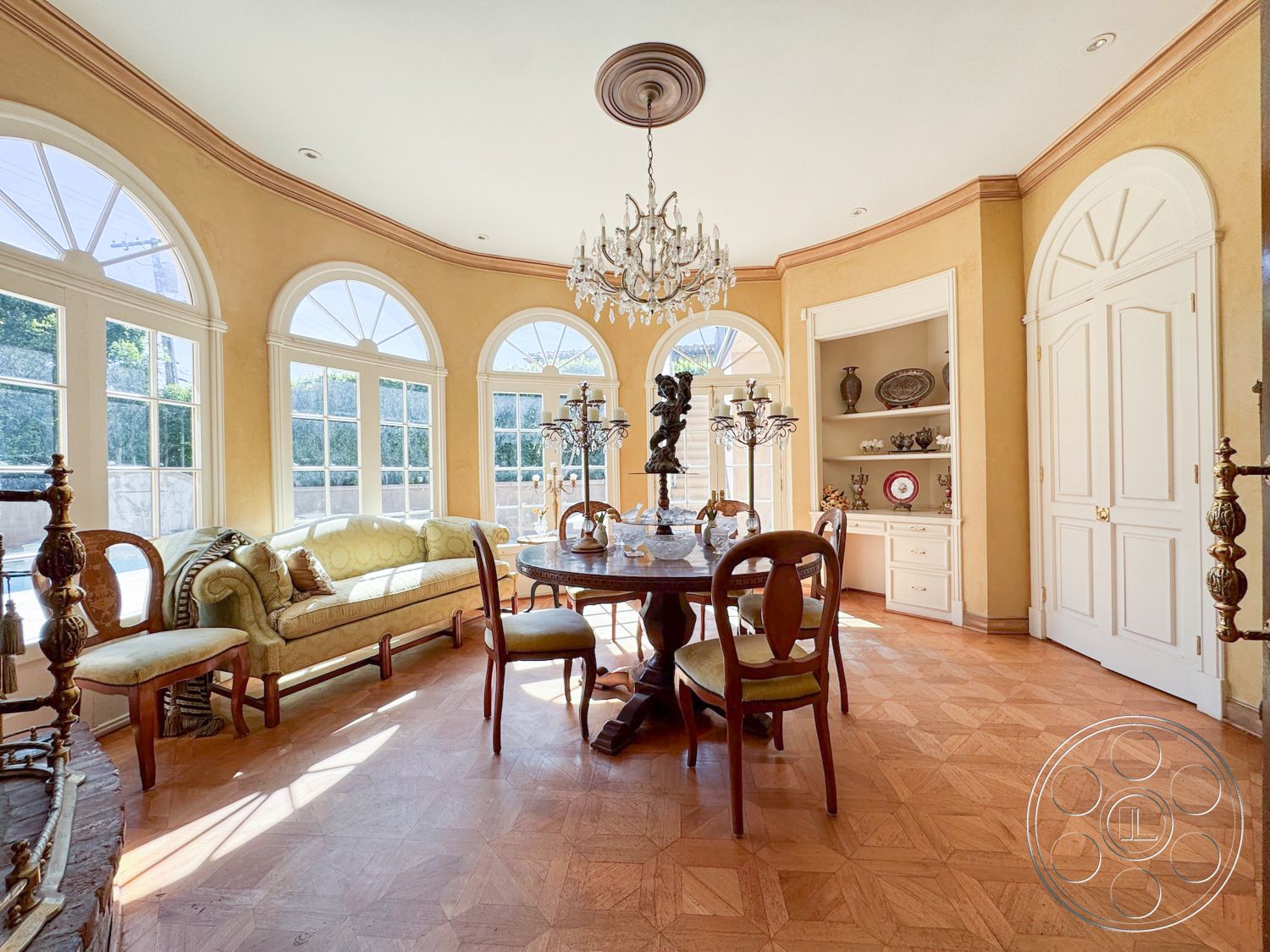 Mansion 146 - round dining table, chandelier lighting, elegant decor accents, arched windows, large window view, interior dining room, classic interior design, crown molding details, parquet wood flooring, natural light ambiance, traditional furniture style, built-in wall shelves, golden yellow wall paint, upholstered sofa with throw pillows, ornate candelabras