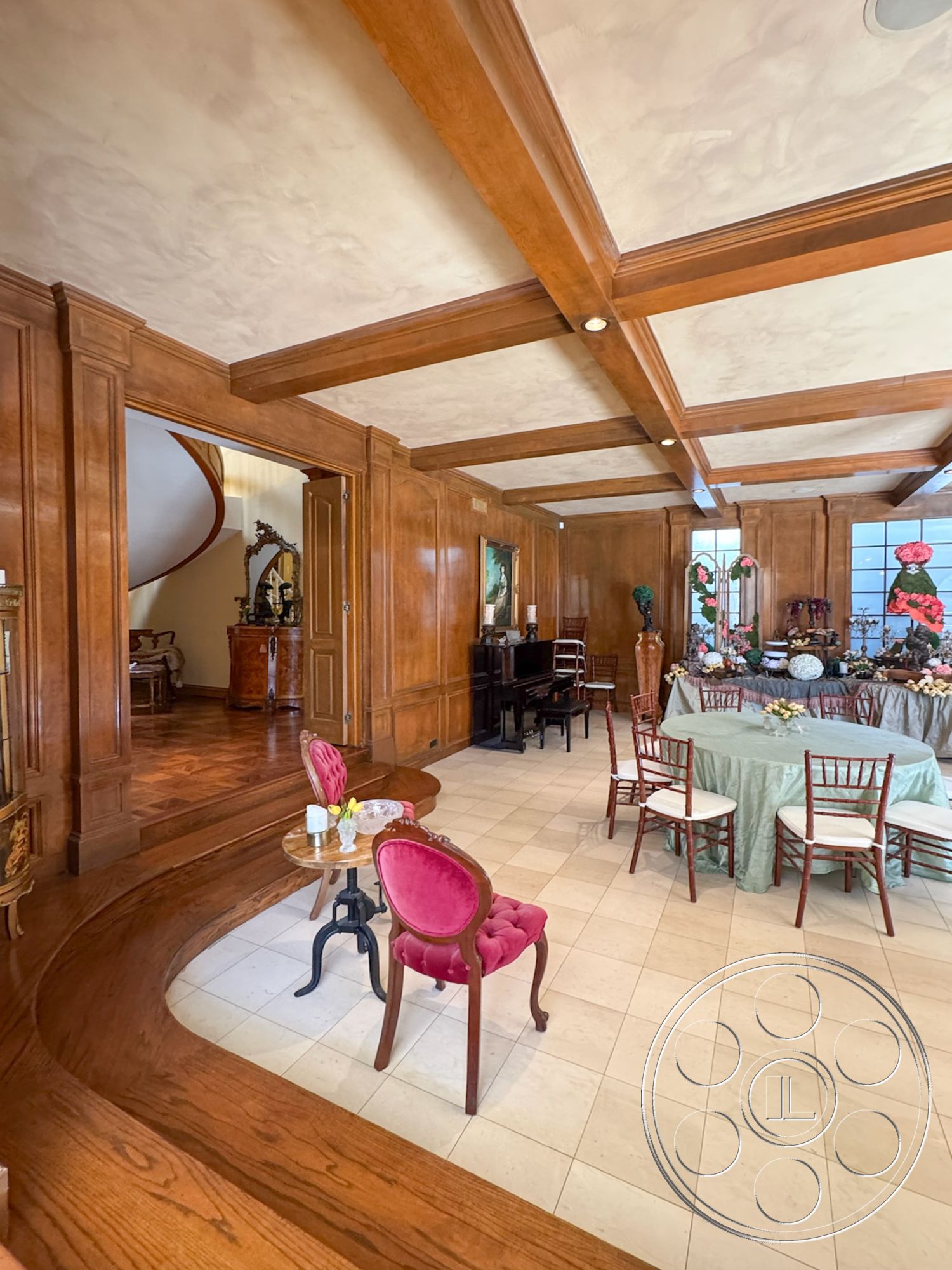 Mansion 146 - textured plaster ceiling, vintage decor elements, natural light from windows, dark wood paneling, exposed wood ceiling beams, traditional furniture style, decorative artwork on walls, hardwood floor dining area, piano in living area, white ceramic tile flooring, round dining table with tablecloth, pink upholstered dining chairs, curved wood transition step, elegantly set dining table, formal dining space