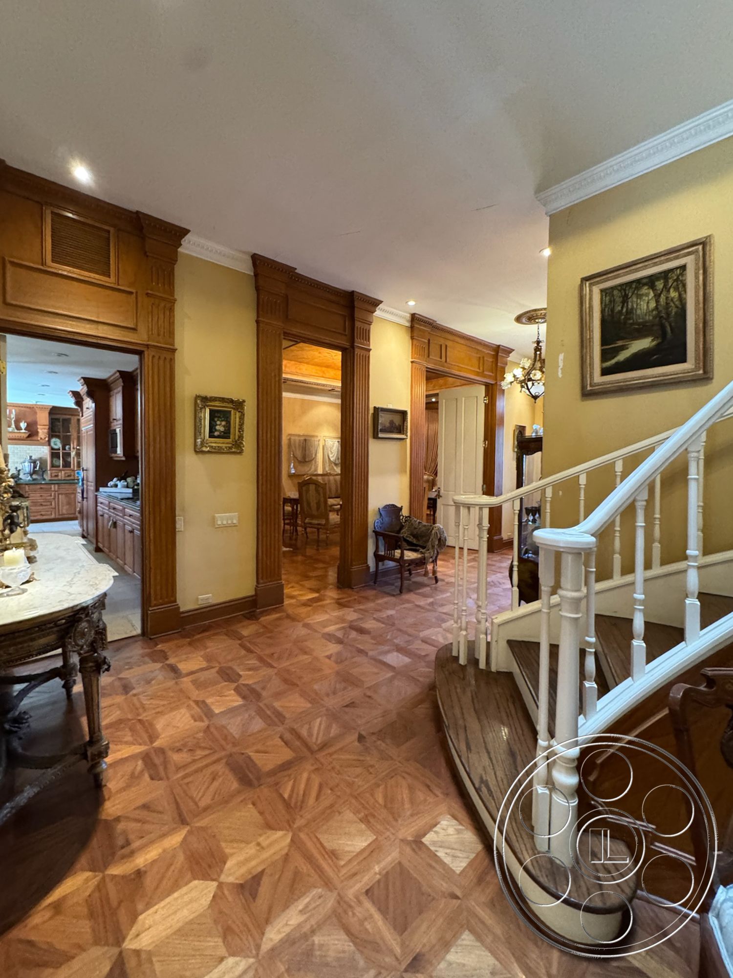 Mansion 146 - open concept kitchen view, hardwood entryway flooring, intricate parquet pattern flooring, classic wooden staircase with curved steps, exposed wood banister with white spindles, golden yellow painted walls, ornate crown molding details, traditional wall art frames, historical dining area access, decorative wrought iron furniture, vintage chandelier lighting, classical room proportions, interior hallway with architectural columns, cohesive design with antique touches, subtle ambient ceiling lighting, light-filled entrance hall, elegant sitting area beside staircase