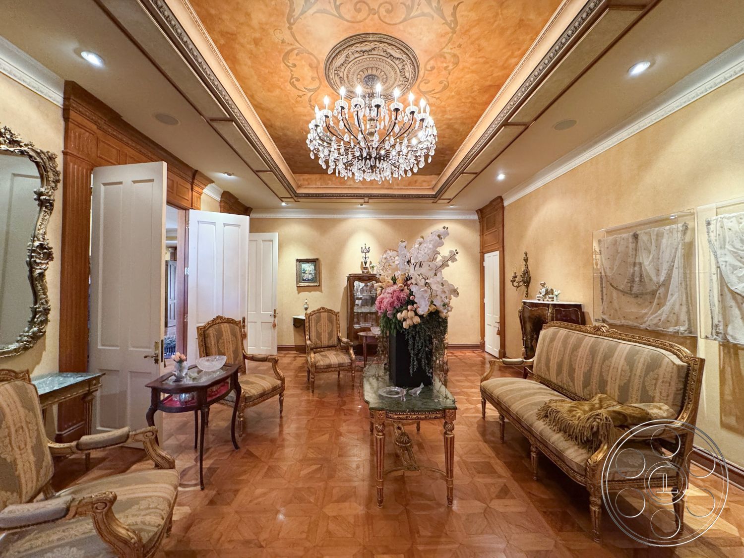 Mansion 146 - decorative crown molding, wood-paneled walls, interior living room, ornate crystal chandelier, natural light from open doors, richly textured wall treatments, stylish accent table, intricate coffered ceiling, plush patterned upholstery, geometric parquet flooring, elegant antique armchairs, golden leaf detailing, classic framed artwork, luxurious floral centerpiece, traditional salon style, symmetrical room arrangements, exotic wood trim, lavish decorative accents