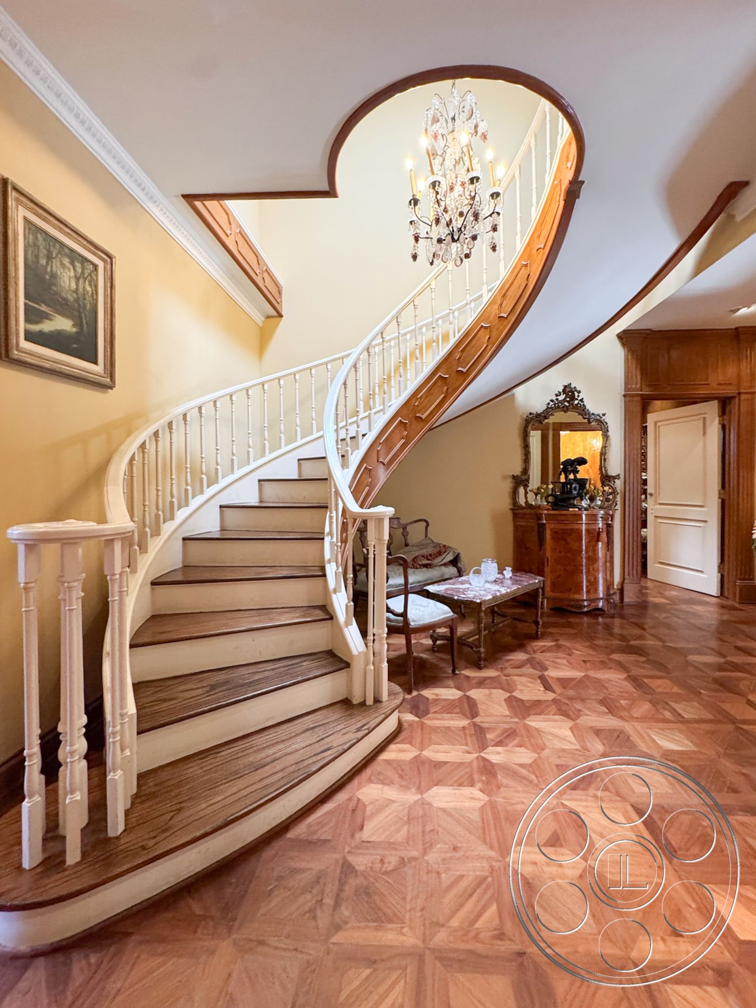Mansion 146 - traditional style home, sculptural staircase design, textured yellow painted walls, curved staircase with wooden railings, parquet hardwood flooring in entryway, chandelier lighting in foyer, exquisite crown molding details, framed landscape art on wall, ornate wood trim around doorways, elegant entryway seating area, open foyer with stair landing, stylish coffee table in seating area