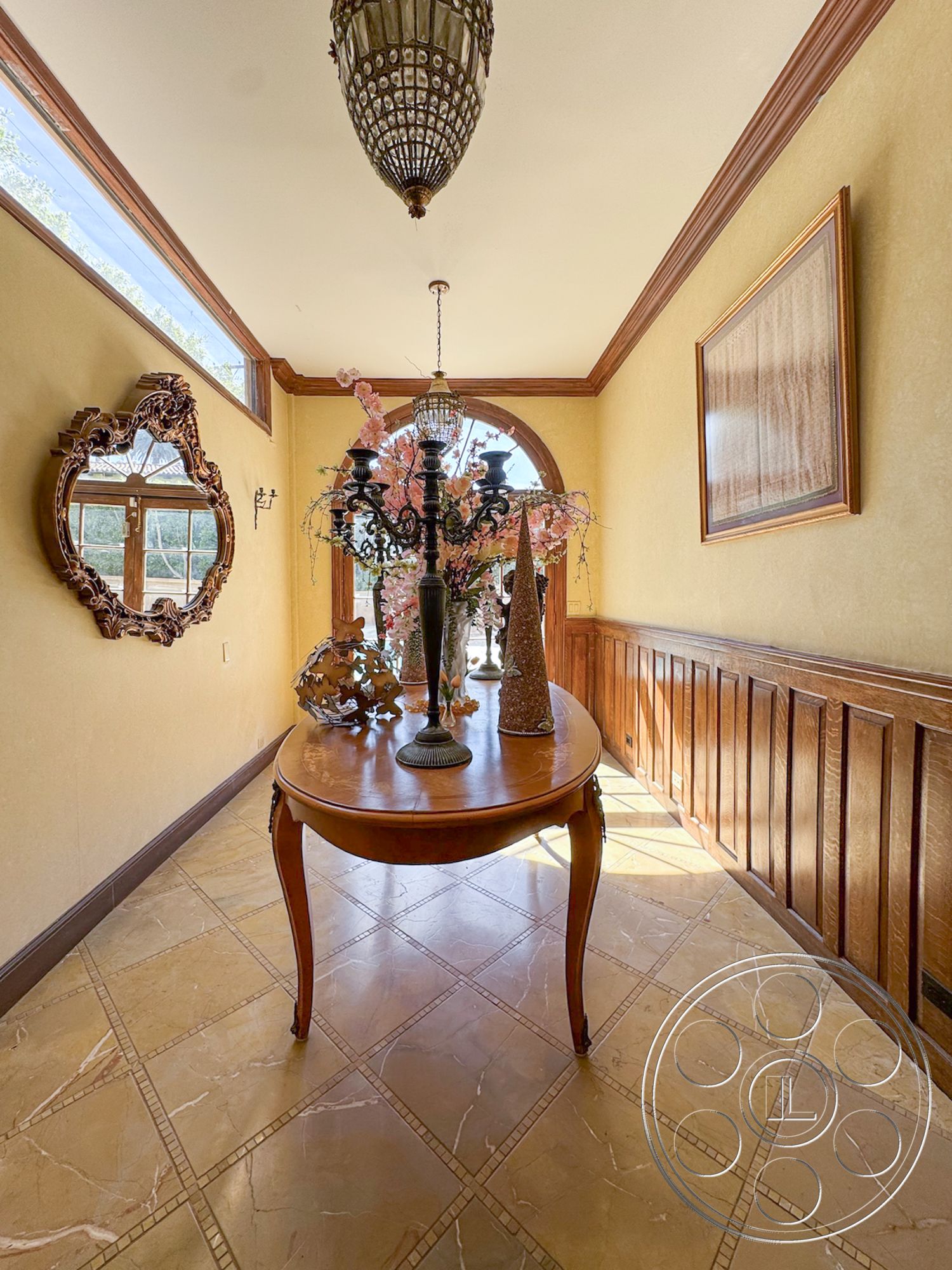 Mansion 146 - interior hallway, decorative floral arrangement, dark wood wainscoting, classic framed artwork, marble flooring with mosaic accents, ornate ceiling chandelier, arched window with natural light, traditional round wooden table, golden wall paint, mirror with intricate detailing, beamed ceiling with crown molding