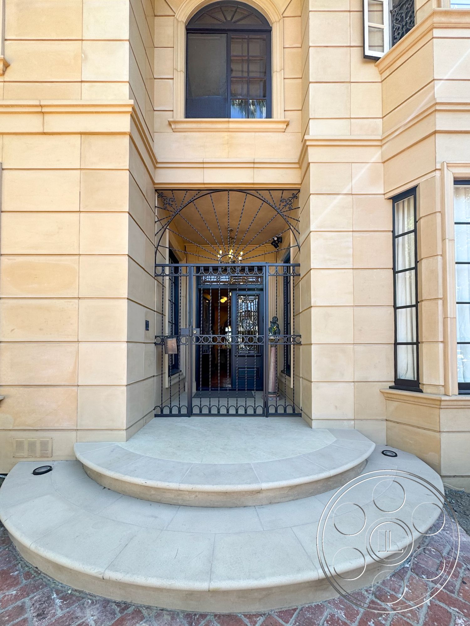 Mansion 146 - arched window, decorative window grilles, wrought iron gate, symmetrical design elements, textured plaster wall accents, traditional architectural details, brick paver walkway, curved stone steps, double-hung window style, exterior grand entrance, sandstone facade walls, elegant entryway lighting, stone lintel, circular entry area, classic home design, ornate wrought iron patterns, suburban luxury residence