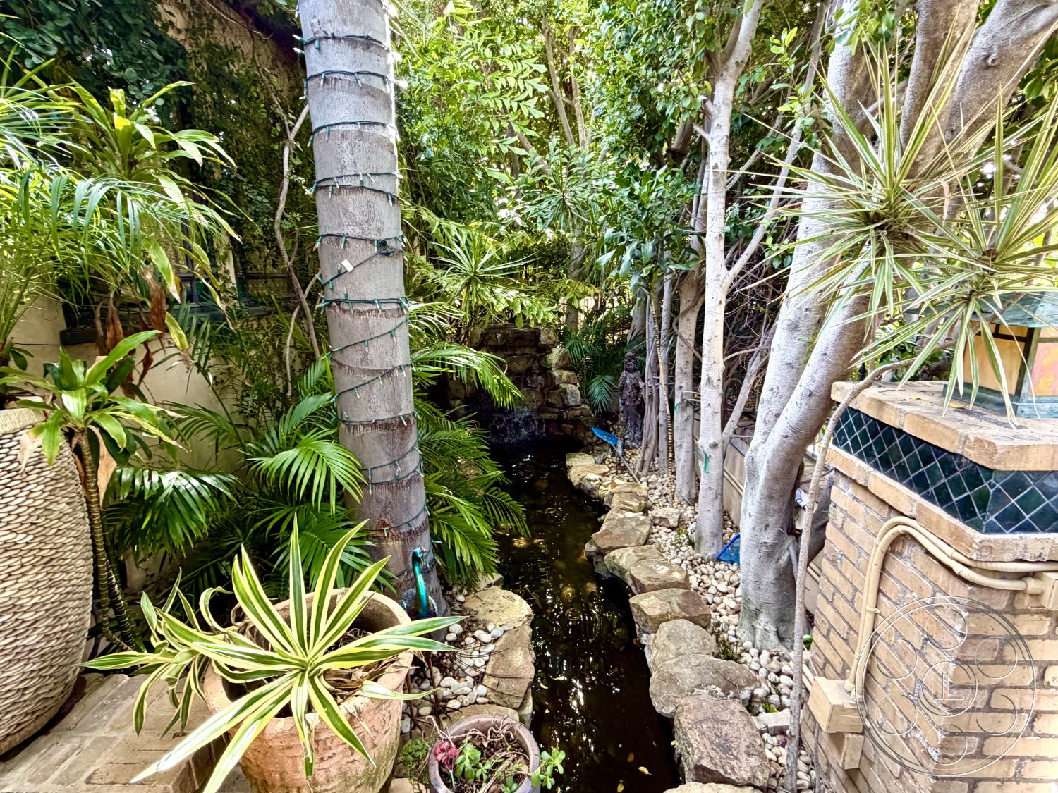Medit 167 - exterior, exposed brick wall, decorative tile accents, natural stone pathway, greenery backdrop, outdoor living area, pebble ground cover, tall tree canopy, tropical garden setting, stone water feature, woven outdoor planter, lush palm landscaping, tranquil creek-like stream, artistic lantern lighting, cozy garden nook