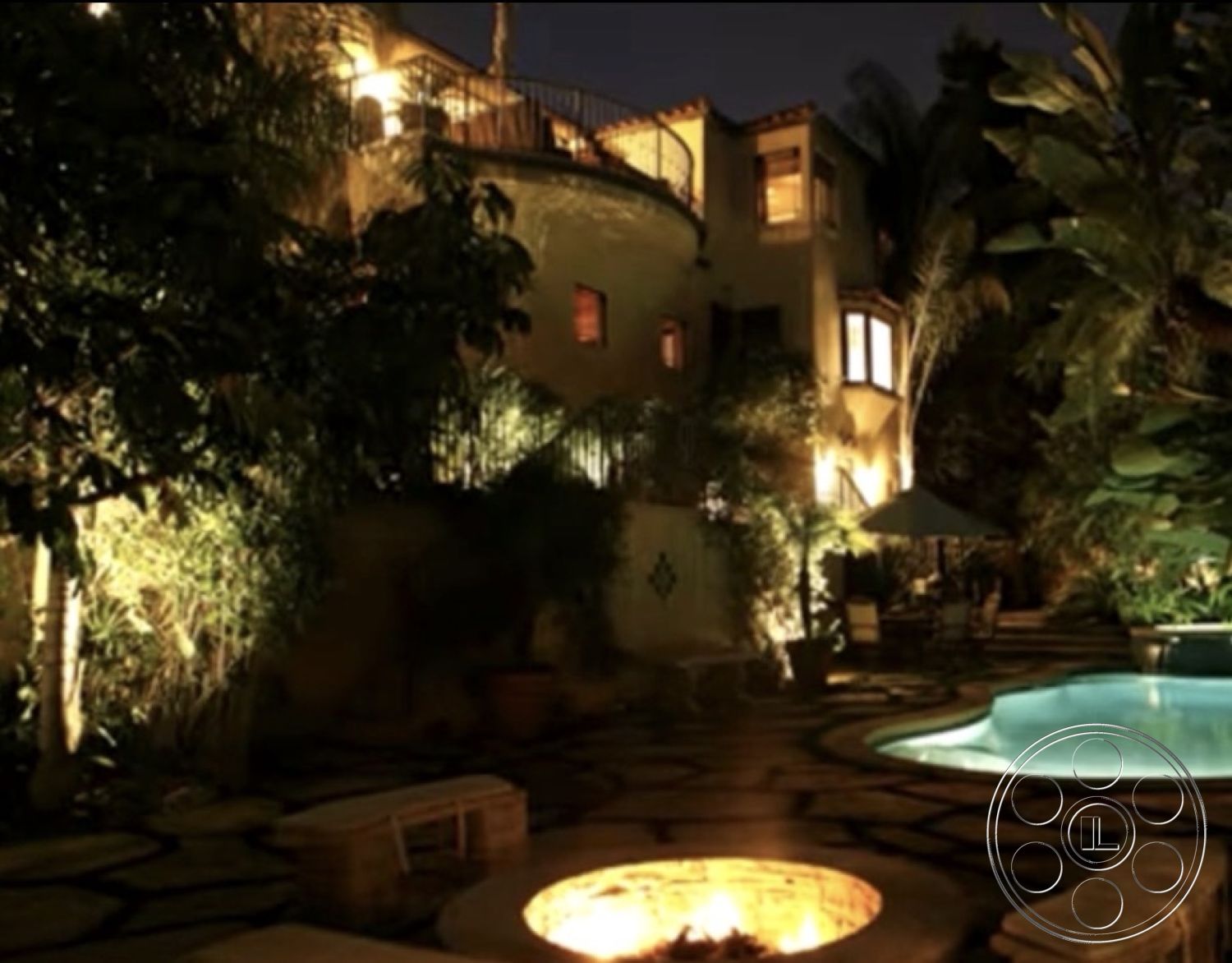 Medit 167 - exterior, palm trees, single-story home, nighttime setting, soft ambient lighting, tropical landscaping, infinity pool, arched windows, Mediterranean architectural style, stucco exterior walls, wooden railing balcony, curved stone patio flooring, exposed stone fire pit, tile rooftop, round patio dining area