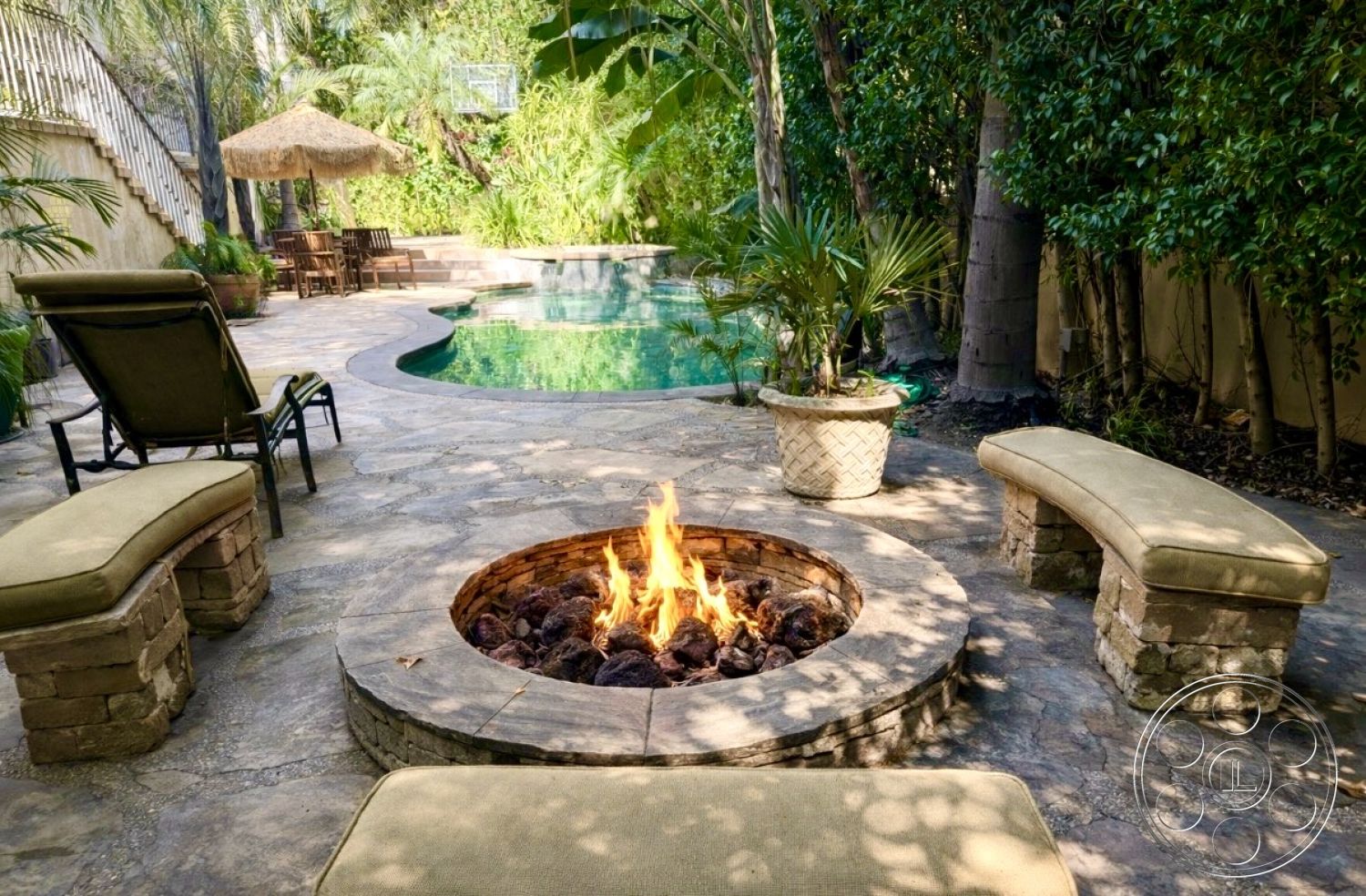 Medit 167 - paved pathway, outdoor lounge chairs, lush greenery backdrop, fire pit area, sunny outdoor space, landscaped garden setting, natural stone patio flooring, exterior pool area, tropical plant accents, decorative planter pots, wooden outdoor dining set, concrete retaining walls, kidney-shaped swimming pool, round stone fire pit, exposed stone fire pit surround, built-in stone seating, soft cushion seating, tranquil poolside atmosphere, umbrella shade structure