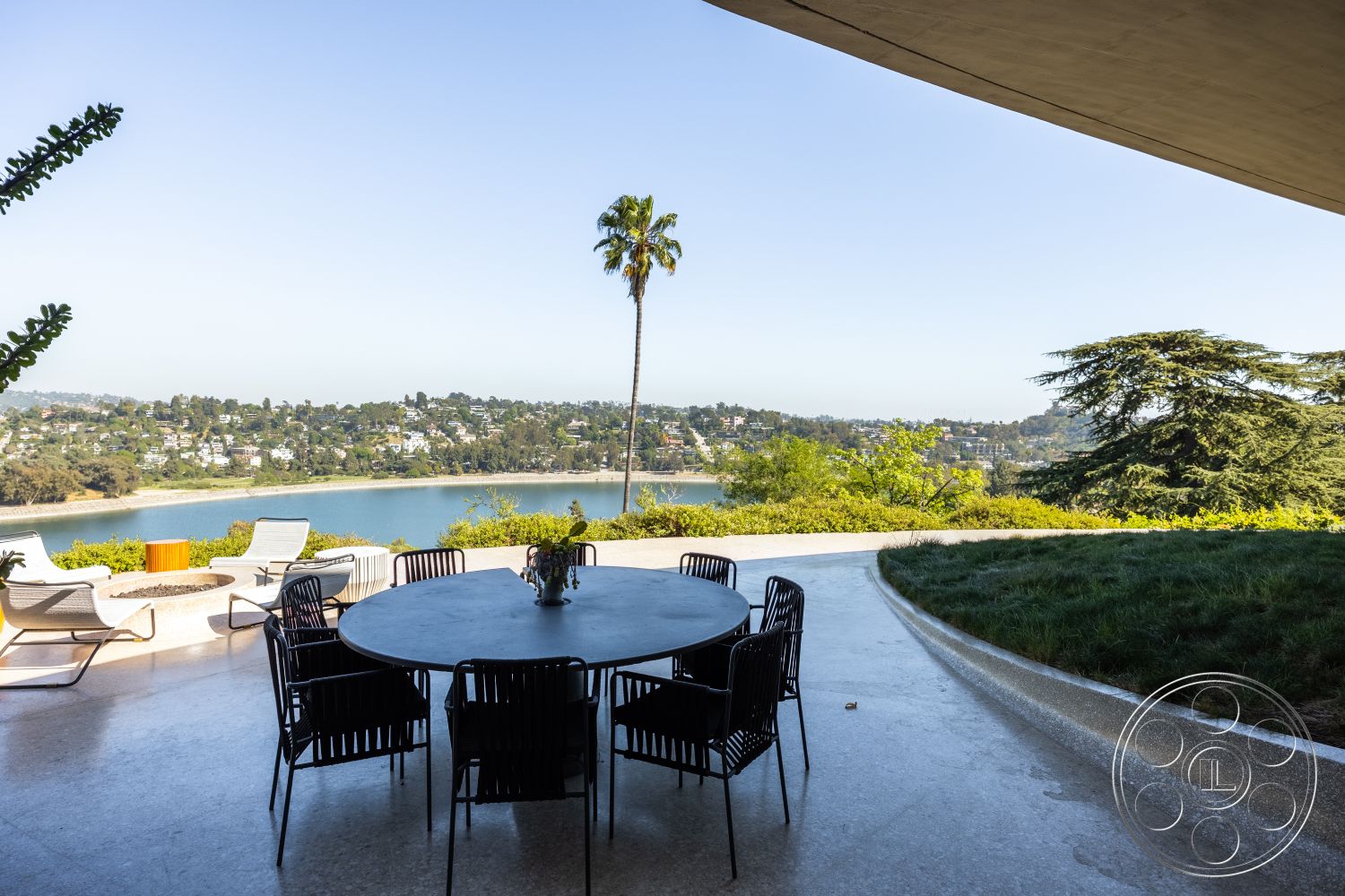 Silvertop House - minimalist design with clean lines, exterior terrace with concrete flooring, circular dining table with metal chairs, modern outdoor seating area with lake view, landscaped garden with grass and shrubs, panoramic view of hillside residential area, palm tree accents in outdoor space, curved pathway leading to lawn, contemporary design elements in outdoor setting, open-air patio with ambient lighting potential, reflective water surface creating serene atmosphere, urban lakeside location, cohesive blend of nature and architecture