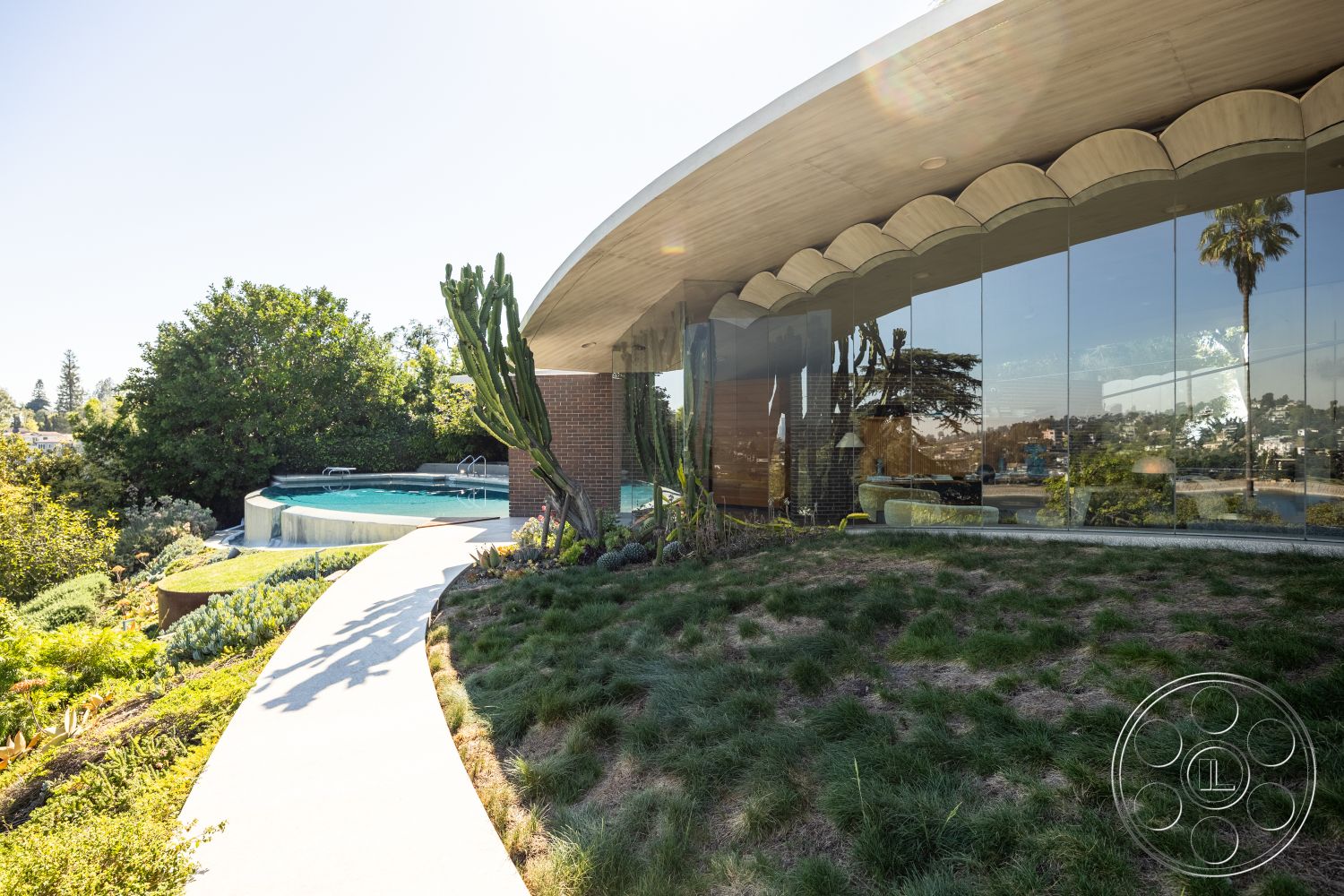 Silvertop House - modern architectural style, large floor-to-ceiling windows, green grass lawn, curved concrete pathway, urban hillside location, concrete patio flooring, concrete pool border, minimalist outdoor design, landscaped garden with native plants, rounded building form, exterior pool area with infinity pool, brick and glass exterior walls, wooden overhanging roof structure, outdoor cactus garden, scenic city skyline view