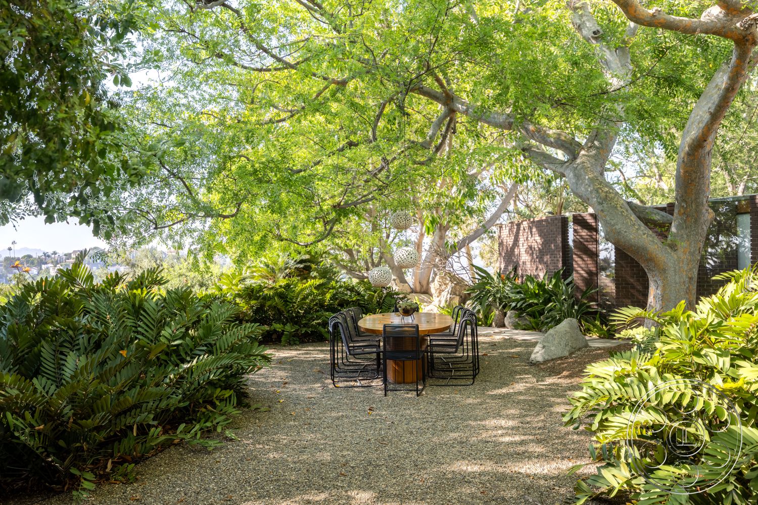 Silvertop House - exterior, wood dining table, tropical plants, outdoor dining area, urban garden, scenic views, garden landscaping, large shade trees, open-air setting, metal dining chairs, gravel patio flooring, lush greenery surrounding patio, rustic outdoor furniture, modern brick wall, boulders as landscape features, peaceful garden retreat