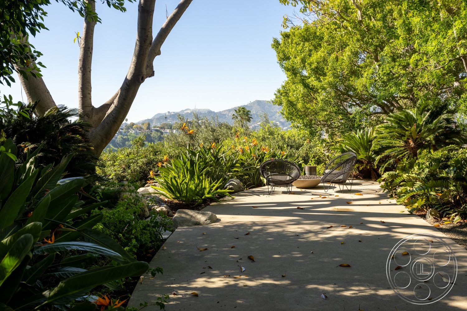 Silvertop House - lush tropical landscaping, diverse plant life, sun-drenched outdoor space, exterior concrete patio flooring, natural stone borders, low-maintenance landscape design, woven patio chairs, scenic mountain view, round fire pit table, shadow play from tree foliage, tranquil outdoor seating area, slightly elevated patio, California-style garden, decorative gravel path, open-air relaxation zone