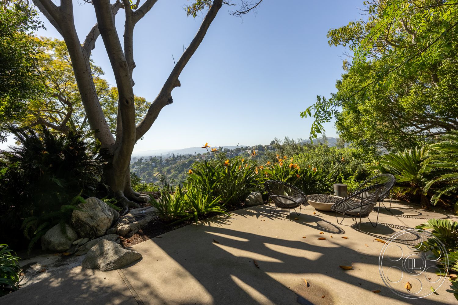 Silvertop House - exterior, outdoor seating area, drought-tolerant landscaping, concrete patio flooring, landscaped garden setting, rock garden elements, sunny hillside location, lush tropical plants, scenic city skyline view, modern patio chairs, fire pit design, planter beds with greenery, partial shade from large trees