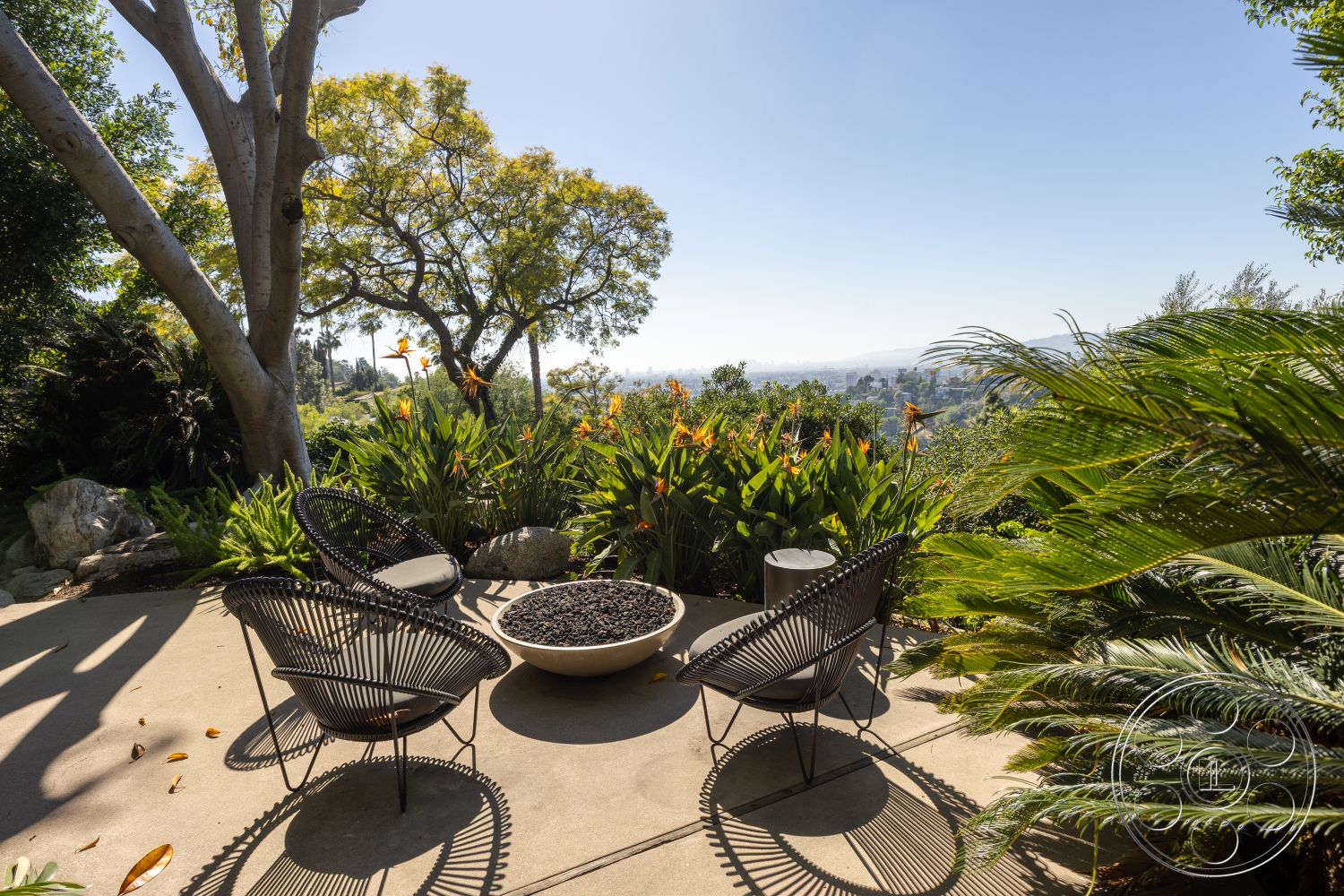 Silvertop House - city skyline view, concrete patio floor, sunlit garden space, tropical garden plants, exterior patio seating area, lush foliage backdrop, natural setting with greenery, modern outdoor fire pit, black wicker lounge chairs, textured stone accent rocks, landscape featuring palm trees, casual outdoor entertainment area, rolling hillside views