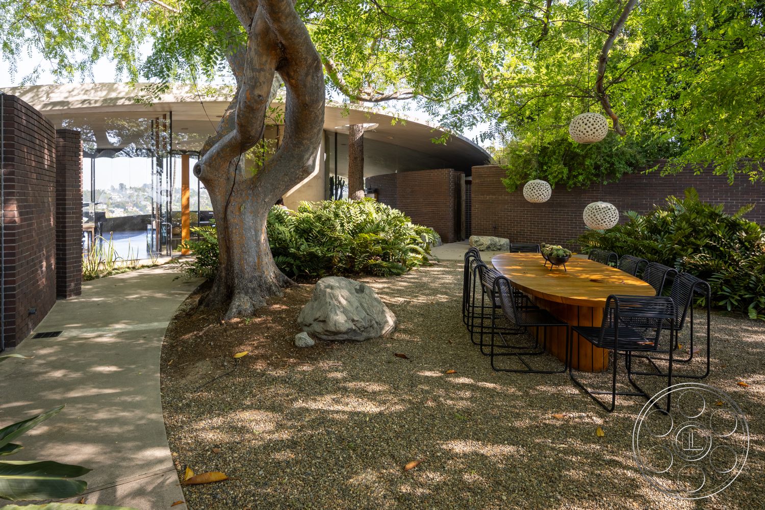 Silvertop House - integrated lighting, outdoor dining area, modern architectural style, round wooden dining table, metal dining chairs, gravel patio flooring, urban garden setting, curved stone pathway, brick wall exterior, hanging decorative light fixtures, lush landscaping with native plants, natural stone rock features, leafy trees providing shade, glass walls for panoramic views, textured natural stone borders