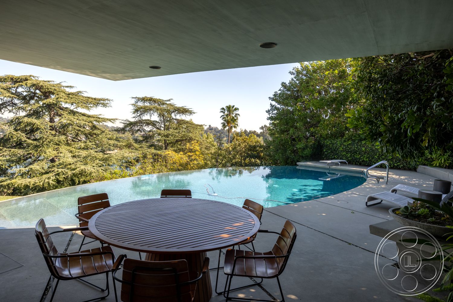 Silvertop House - concrete flooring, secluded backyard, concrete pool deck, lush greenery view, metal outdoor chairs, urban hillside location, contemporary outdoor living space, modern minimalist architecture, sunny outdoor patio, exterior infinity pool, wooden patio dining table, landscaped garden with mature trees, glass railing overlooking landscape, open-air outdoor setting