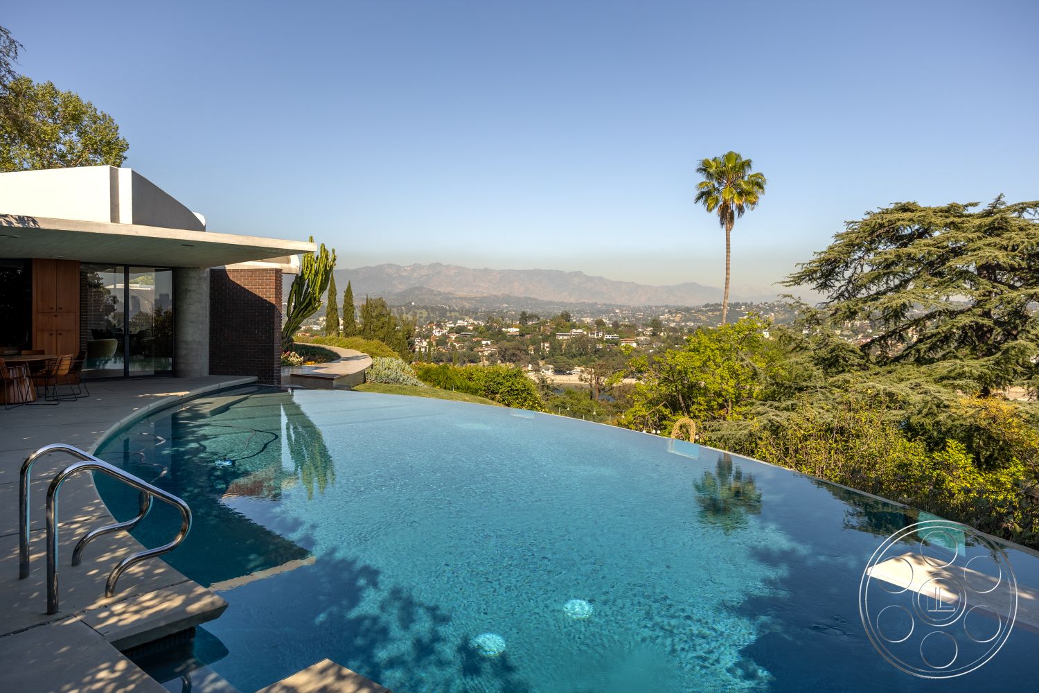 Silvertop House - minimalist design elements, wooden dining table, outdoor seating area, infinity pool, open-air living space, tropical palm trees, stone pool deck, contemporary architectural style, landscaped garden area, urban hillside location, surrounding mountain views, exposed concrete patio floor, sunny exterior setting, large floor-to-ceiling glass windows, modern wooden deck railing