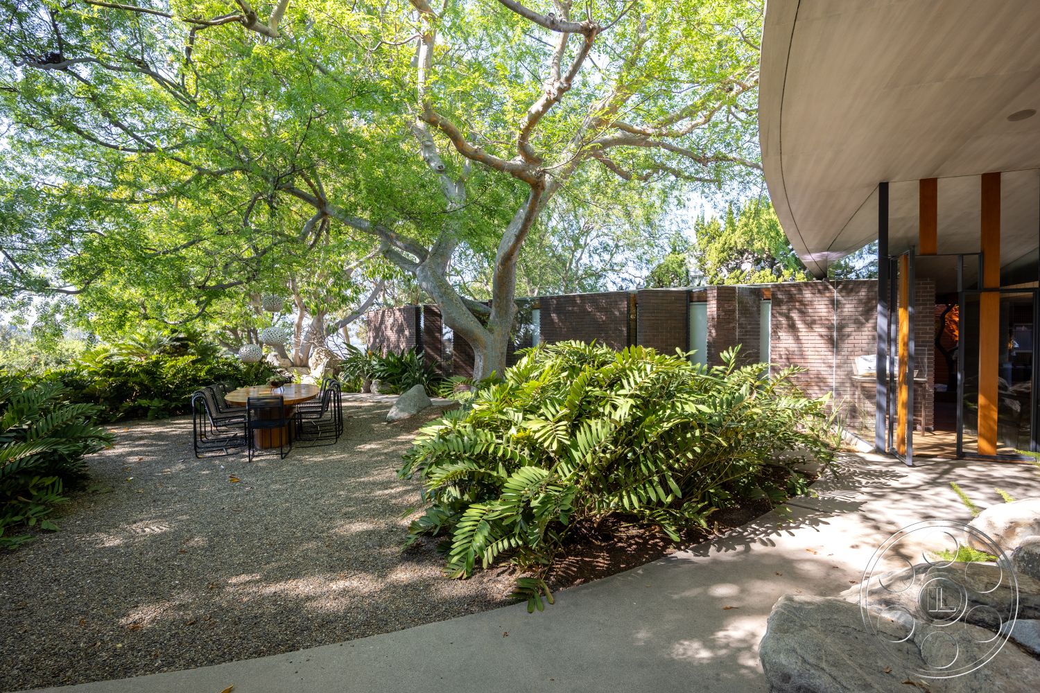 Silvertop House - wood dining table, exposed brick walls, floor-to-ceiling windows, outdoor dining area, lush landscaping, large shade trees, mid-century modern architecture, curved rooflines, open-air layout, hardscape features, metal dining chairs, gravel garden path, urban garden setting, natural stone boulders, concrete patio floor, native ferns, interior outdoor integration
