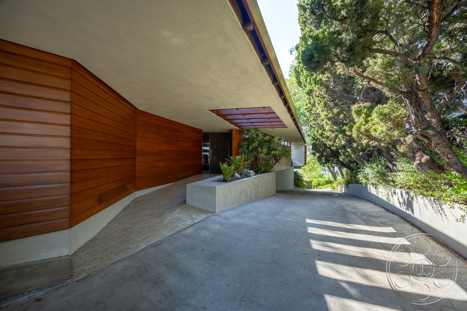 Silvertop House - low-maintenance landscaping, smooth concrete surface, modern architectural style, natural wood tones, horizontal wood slats, exterior concrete driveway, exterior landscaped garden, concrete planter box, natural light emphasis, exterior wood paneling, angled roof design, textured stucco wall, minimalist entryway, tropical garden setting, shadow play from trees, curved driveway path, built-in greenery feature