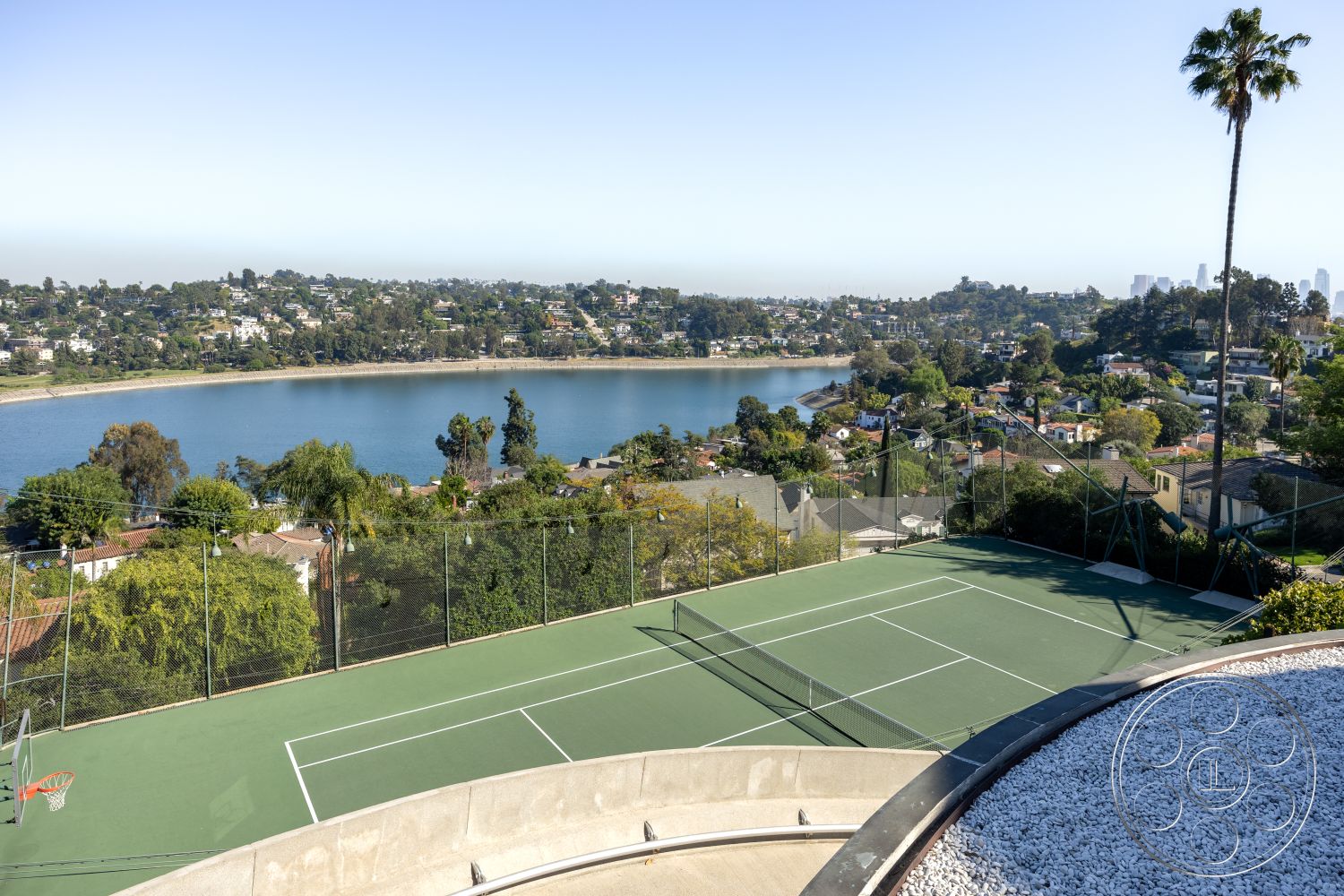 Silvertop House - landscaped hillside, stone retaining wall, outdoor entertainment area, tropical palm tree landscaping, open-air recreational space, urban skyline background, fenced tennis court, gravel patio area, concrete tennis court surface, expansive lakefront view, sloped terrain tennis court, curved outdoor terrace, blooming garden elements, interior exterior transition space, modern hillside property