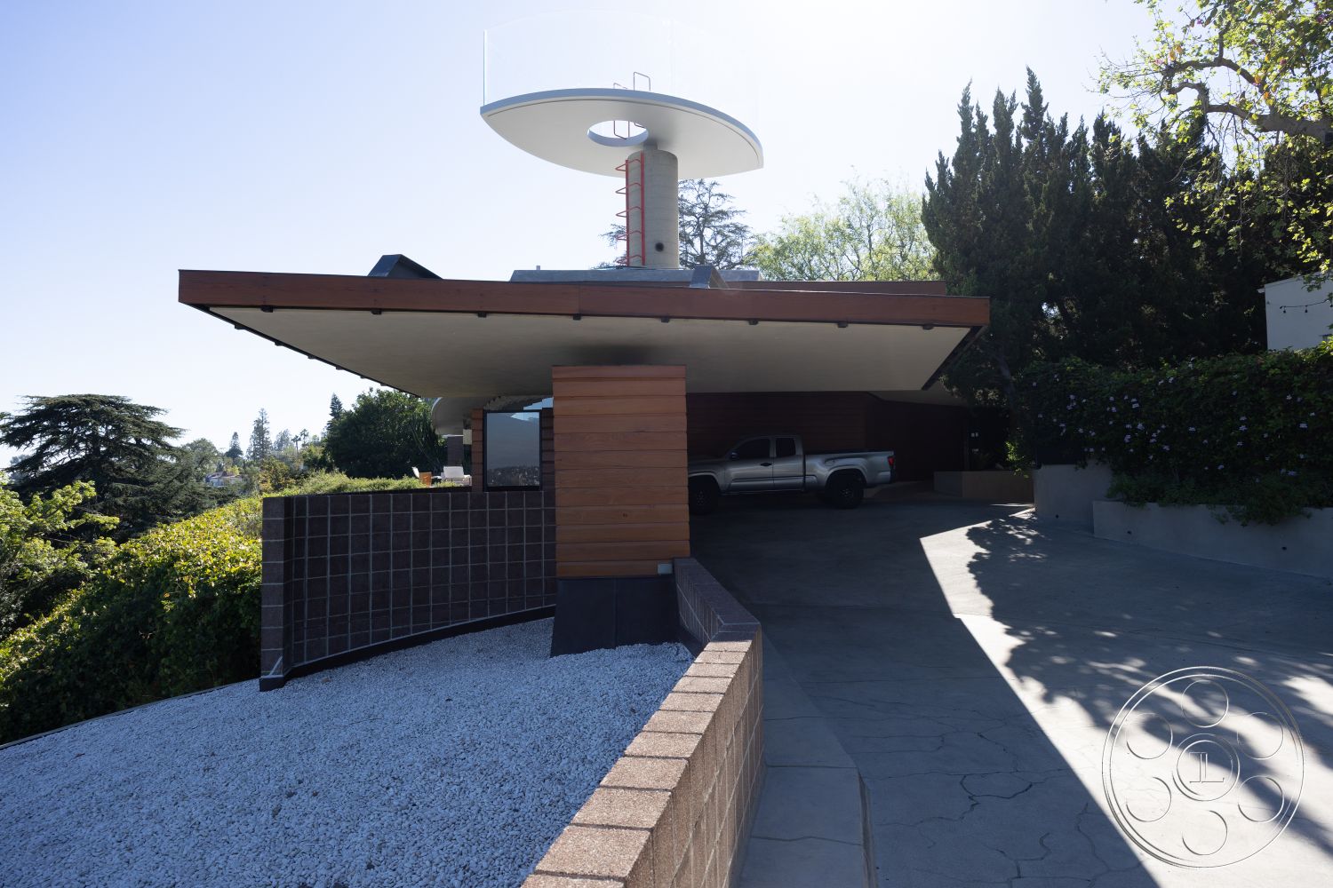 Silvertop House - exterior concrete driveway, exterior landscaped garden, exterior gravel landscaping, exterior minimalist aesthetic, setting hillside location, exterior glass balcony railing, architectural modern design, exterior smooth stucco finish, exterior wooden cladding, exterior overhanging roof structure, exterior brick retaining wall, structural cantilevered roof, shaped lawn with hedges, exterior circular roof feature, setting urban neighborhood, setting wooded backdrop