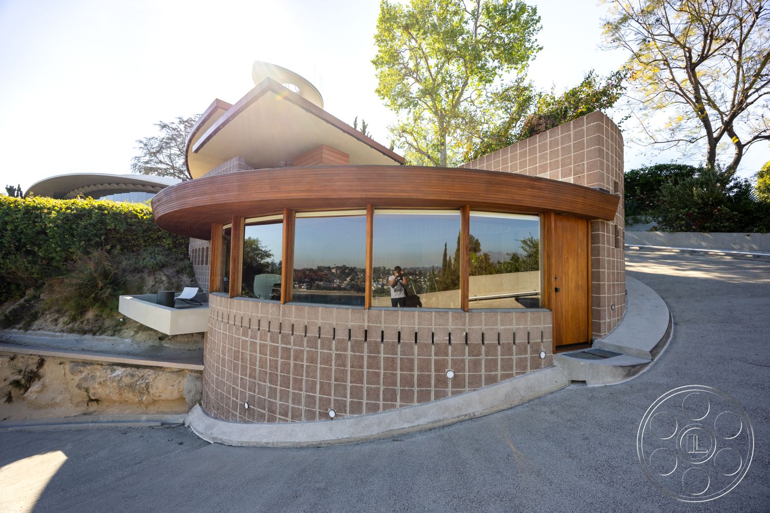 Silvertop House - exterior, contemporary design, modern architectural style, large floor-to-ceiling windows, geometric forms, natural wood front door, hillside location, smooth concrete driveway, sunlit exterior, surrounding green landscaping, light-colored concrete block walls, curved wooden roof overhang, rounded architectural elements, woven wood detailing, open view of the surroundings