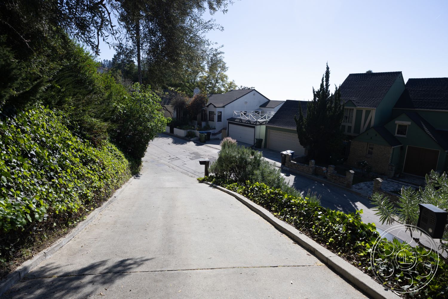 Silvertop House - concrete driveway, curved driveway, surrounded by greenery, urban residential area, textured concrete surface, sunny hillside location, landscaped hillside setting, exterior stone accents, multi-story homes, wooden fence details, plant-lined driveway