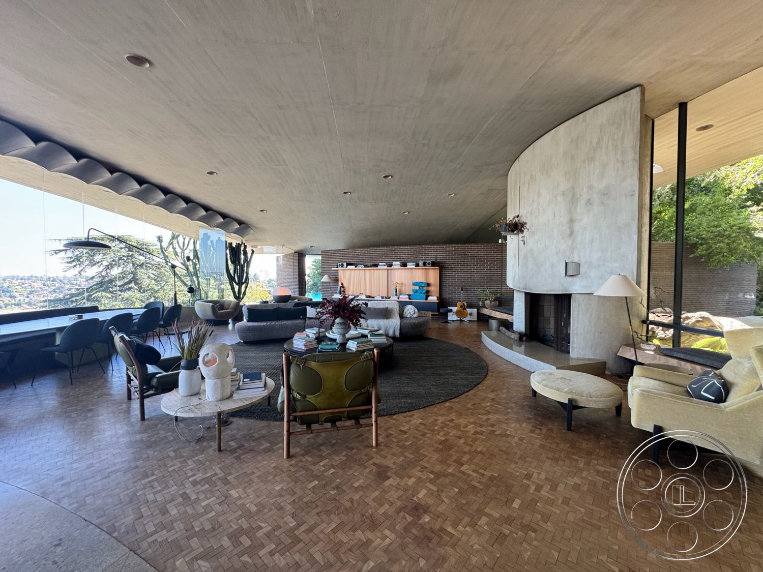 Silvertop House - neutral color palette, cozy seating arrangement, seamless indoor-outdoor transition, vaulted ceiling, brick accent wall, open concept living space, exposed concrete ceiling, round area rug, indoor plant decor, mid-century modern architectural style, natural light through large windows, modern accent chairs, glass wall with city skyline view, minimalist lighting fixtures, built-in wood cabinetry, modern coffee table design, interior parquet wood flooring, curved concrete fireplace, textured fabric sofas, sculptural decor objects