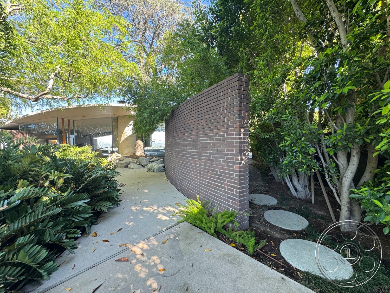 Silvertop House - exterior, large glass windows, outdoor seating area, open-air patio, stepping stone path, smooth concrete pathway, landscaped garden area, modern architectural design, sustainable landscape design, curved brick wall, integrated natural elements, stone accent boulders, lush greenery with shrubs