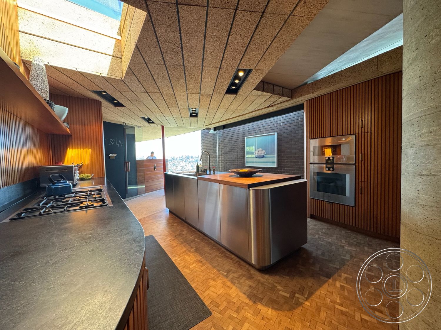 Silvertop House - open concept layout, city skyline view, black stone countertop, interior kitchen, built-in kitchen cabinets, skylight ceiling, parquet hardwood flooring, wood paneling walls, stainless steel kitchen island, cork ceiling tiles, exposed concrete column, modern gas cooktop, brick backsplash, sunlit kitchen space