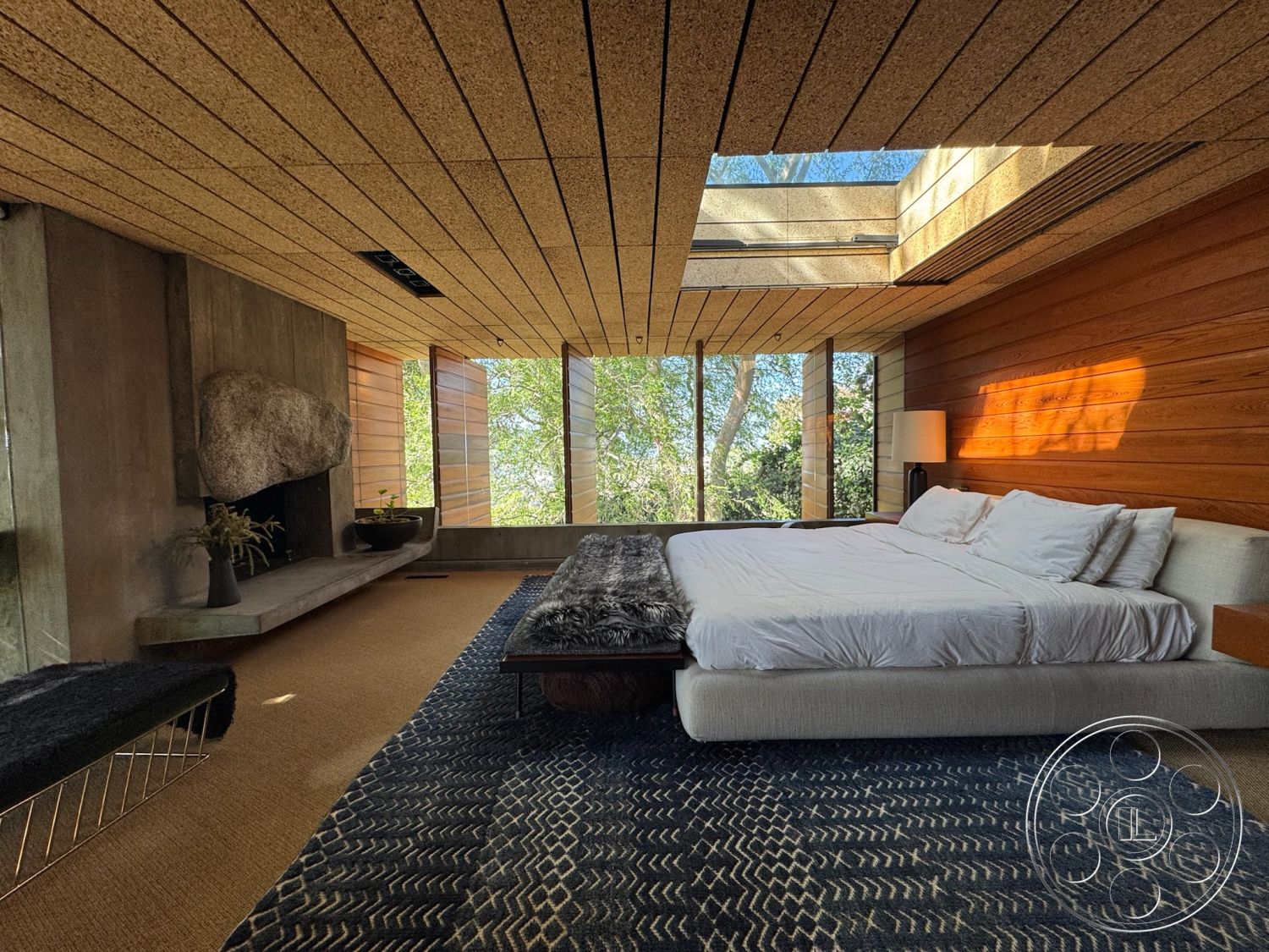 Silvertop House - sliding glass windows, contemporary interior design, natural light-filled space, wood paneling walls, flush mount ceiling lights, king-size bed with white bedding, soft area rug with geometric pattern, exposed concrete fireplace wall, modern minimalism style, vaulted ceiling with wood slats, built-in seating with wood frame, skylight above bed, brown wood accent wall, curated indoor plants