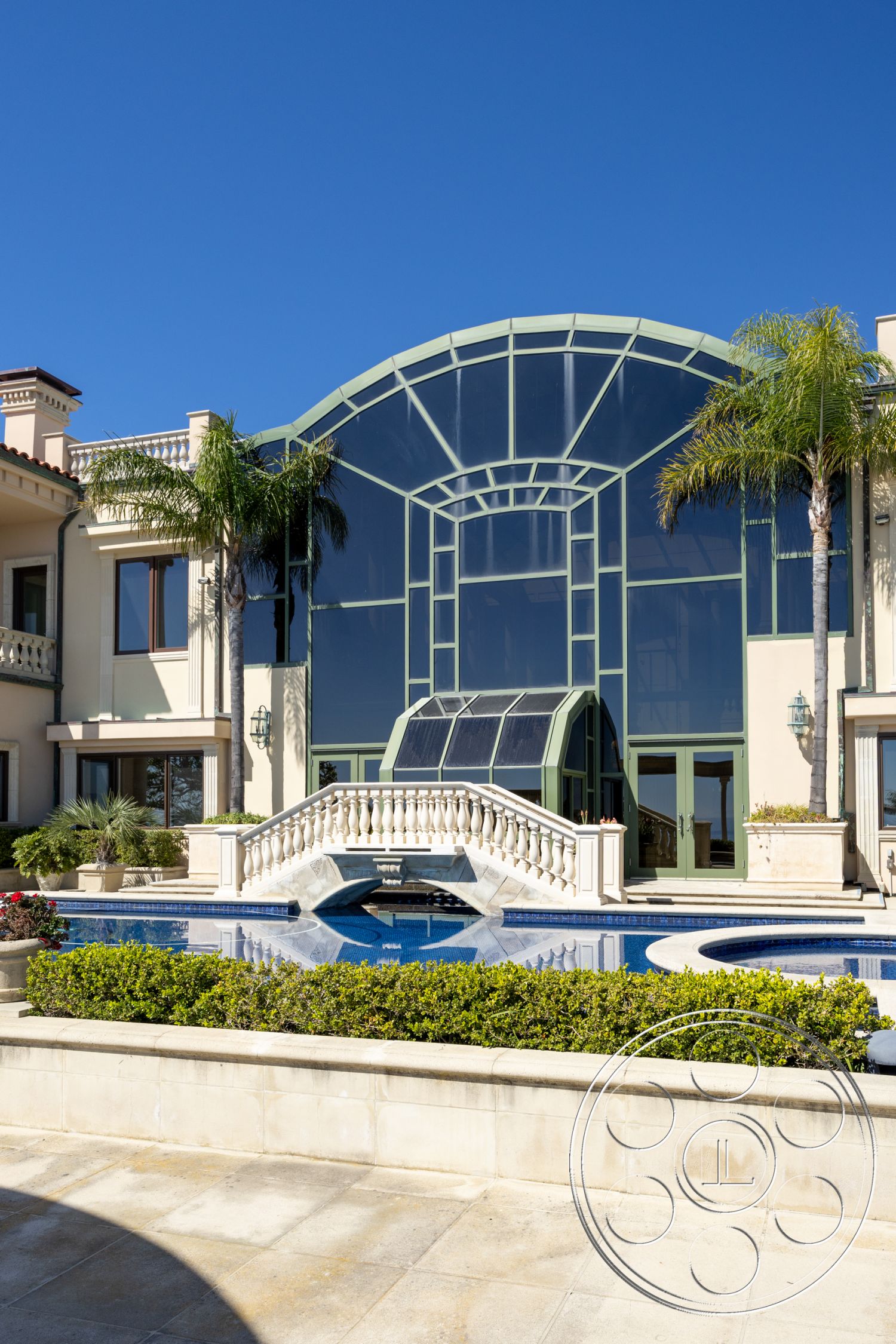 Pacifica Estate - outdoor living space, large glass windows, landscaped gardens, stucco walls, rectangular swimming pool, concrete pool deck, modern architectural style, open-air courtyard, sunny coastal setting, high vaulted ceilings, exterior glass-panel façade, arched glass entrance, cast stone balcony railing, decorative bridge over pool, tropical palm trees landscaping, blue tile swimming pool, imported tile patio flooring, elegant exterior lighting fixtures, paved pathway access, water feature around pool