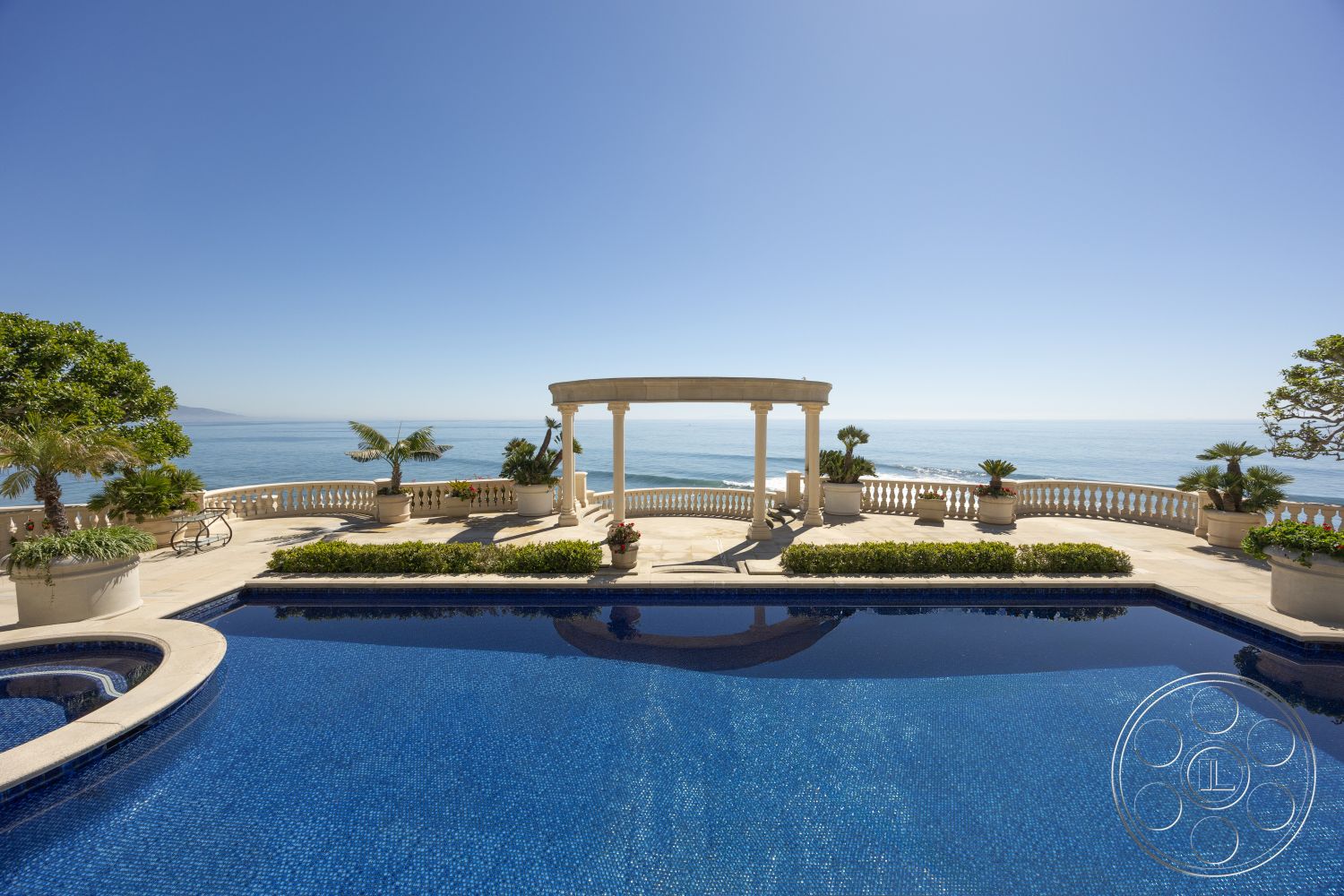 Pacifica Estate - infinity pool, spacious outdoor area, curved pool design, Mediterranean architectural style, stone patio flooring, luxurious outdoor living space, tile pool deck, concrete patio border, landscaped garden with potted plants, oceanfront setting, open outdoor terrace, exterior columns, view of the ocean, sunny coastal environment, decorative railings, plant-filled garden area, understated elegance in design