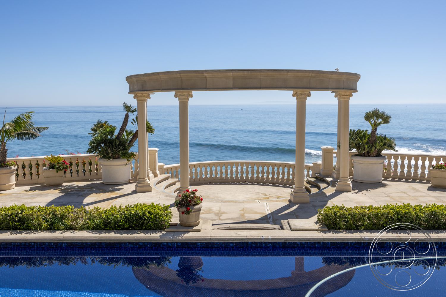 Pacifica Estate - outdoor seating area, stone patio flooring, decorative planter pots, coastal oceanfront view, blue tile swimming pool, rounded stone colonnade, landscape garden with palm trees, curved terrace railing, exterior colonnade, sunny seaside setting