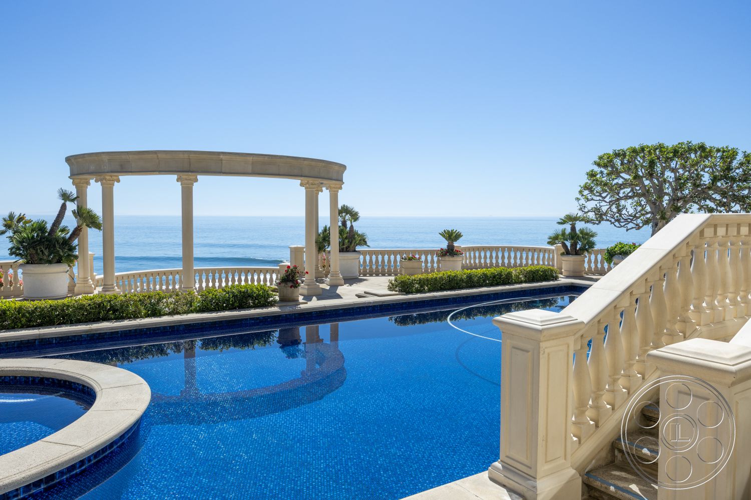 Pacifica Estate - sunny coastal setting, tropical palm trees, modern outdoor seating area, exterior oceanfront property, lush garden landscaping, classical architectural style, rectangular swimming pool with mosaic tile, limestone pool deck, rounded pool border, ornamental columned pergola, staircase with decorative balustrades, paved patio with embedded plant beds, clear blue sky with ocean view, natural stone steps leading to pool, smooth concrete edge for pool, floral planter arrangements