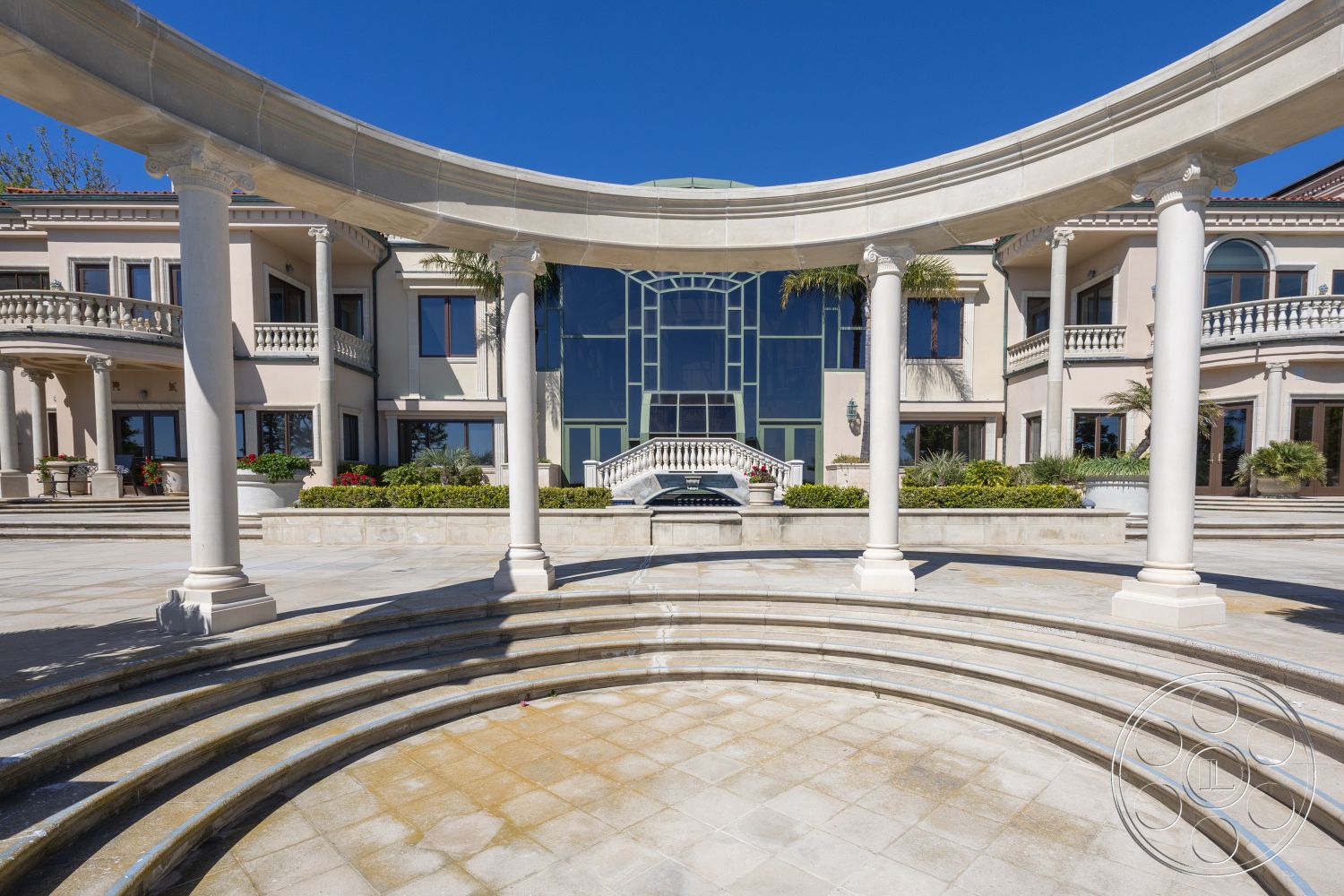 Pacifica Estate - exterior, large glass sliding doors, landscaped surroundings, sunny outdoor space, modern Mediterranean style, stone patio flooring, planted garden area, arched entryway design, ornate column details, curved colonnade structure, two-story luxury home, upstairs balcony with railings, polished marble steps, expansive outdoor layout