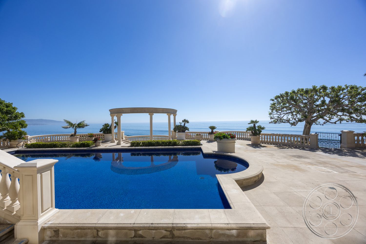 Pacifica Estate - open-air living space, sunny coastal setting, exterior patio with travertine stone flooring, rectangular infinity pool with deep blue tiles, classical Roman-style gazebo with columns, lush tropical landscape with palm trees, oceanfront view overlooking the sea, elevated pool area with decorative stone railings, planter boxes with colorful flowers, staircase with ornate balustrade, outdoor lounge area with gentle sea breeze, surrounding coastal hills, shade provided by mature trees