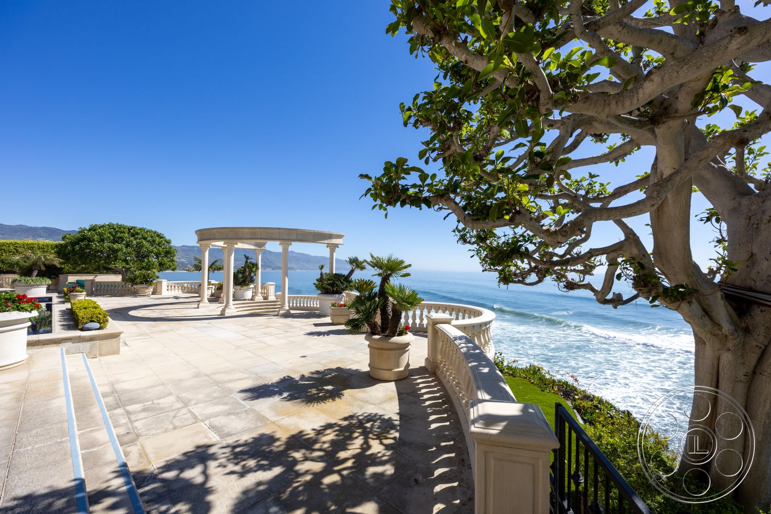 Pacifica Estate - curved walkway, lush tropical landscaping, palm trees landscaping, exterior patio flooring, decorative planter pots, coastal oceanfront view, stone tile patio floor, round column pergola, curved stone railing, blue sky background, sea view terrace, clear sunny day setting, custom outdoor seating area
