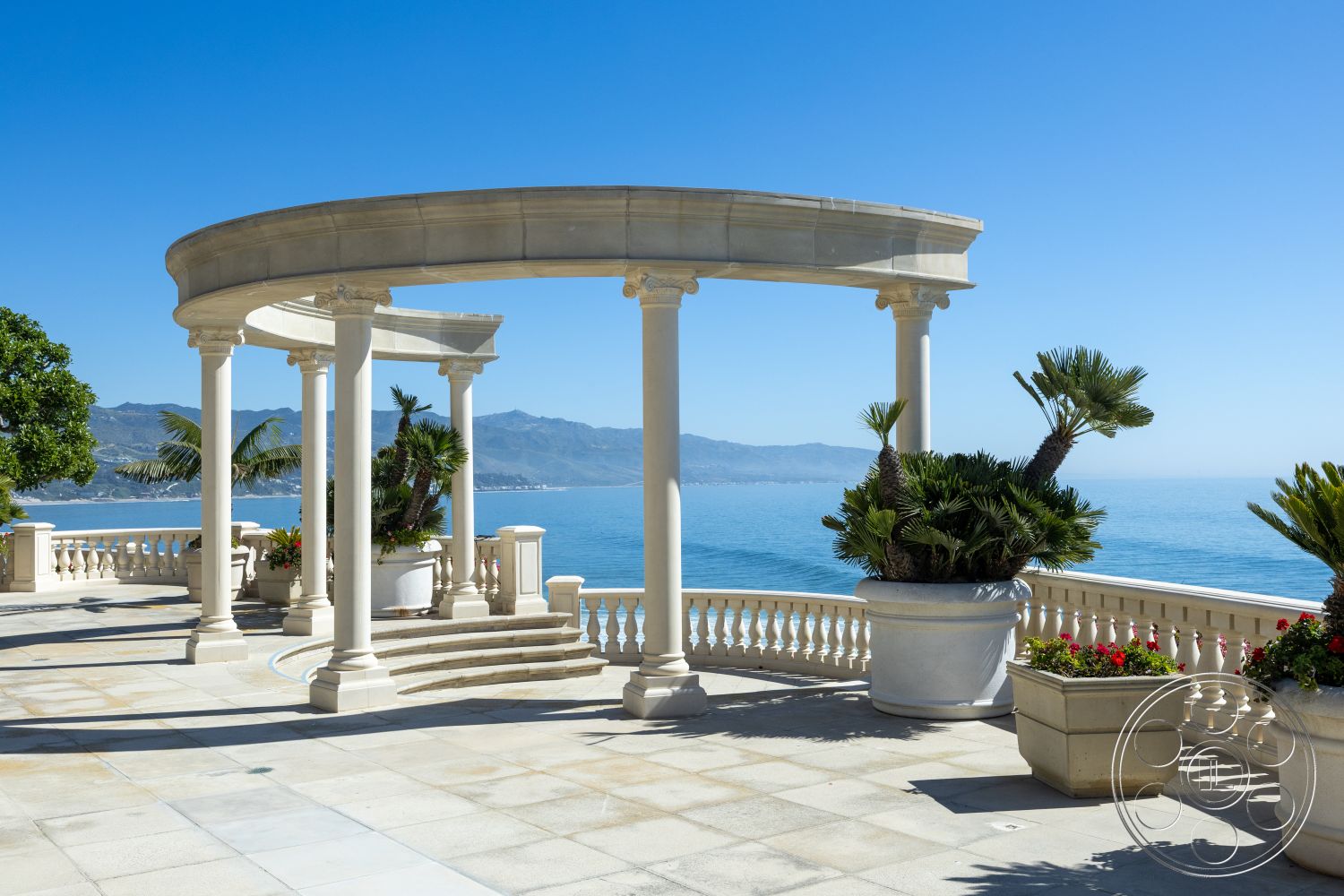 Pacifica Estate - exterior, tropical landscaping, elegant outdoor setting, stone patio flooring, lush palm trees, clear blue sky backdrop, coastal mountain view, open-air architecture, columns with decorative capitals, circular pergola design, oceanfront balcony view, white stone balustrade, rectangular flower planters, sunlit stone flooring, modern outdoor space layout, serene coastal environment, luxury home terrace