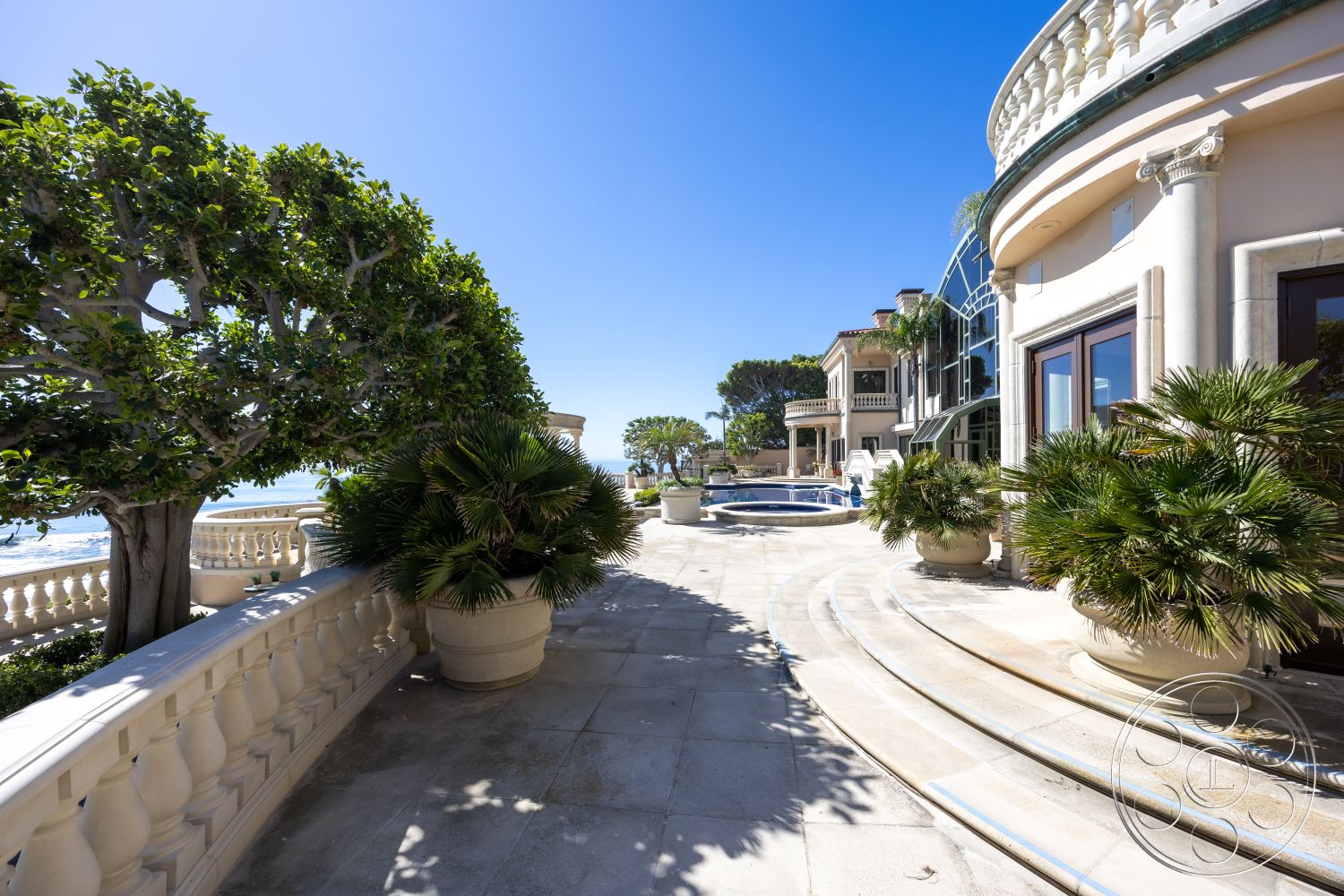 Pacifica Estate - exterior, tropical landscaping, sunny coastal setting, outdoor living area, curved pool design, balustrade balcony, coastal home architecture, exterior glass windows, marble patio flooring, sea view terrace, rounded patio edges, stone railing details, palm tree planters, luxury oceanfront property, elegant facade details