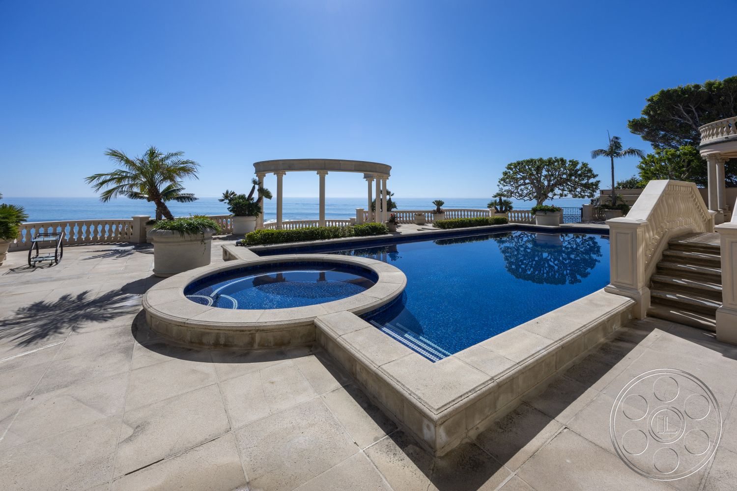 Pacifica Estate - outdoor seating area, rectangular swimming pool, circular hot tub, ocean view, textured stone walls, stone patio flooring, palm trees landscaping, exterior pool area, luxurious outdoor living space, granite pool border, sunny coastal environment, expansive oceanfront view, classical column structure, patio with decorative railings, poolside garden beds, bricked stairway to pool, clean modern lines