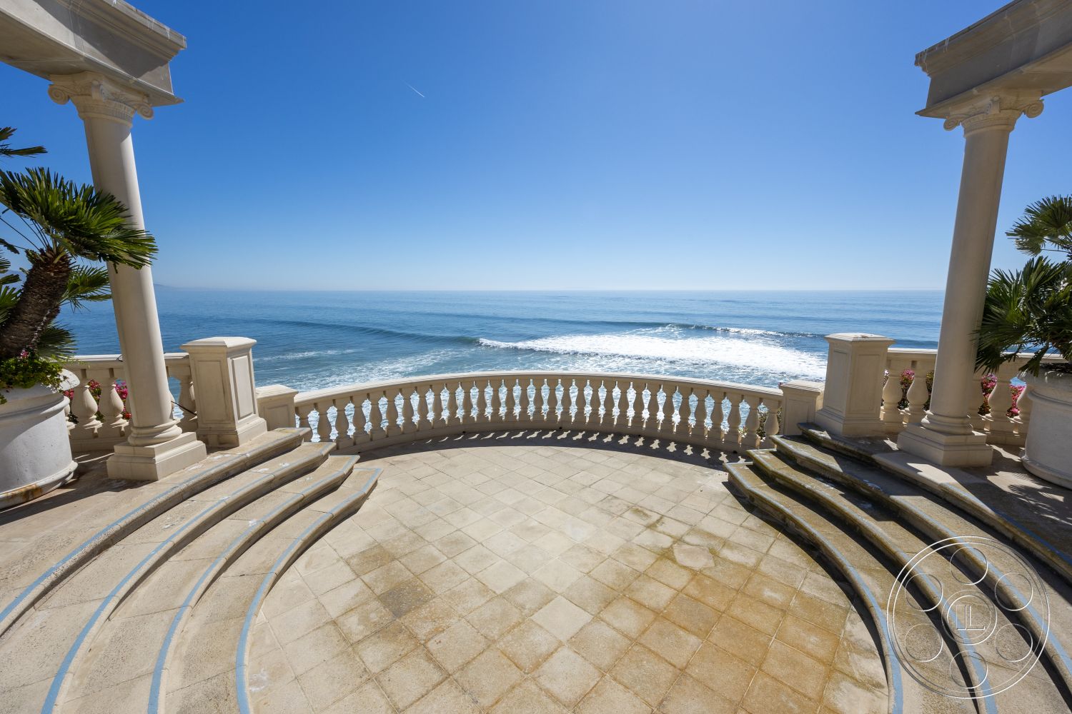 Pacifica Estate - outdoor living area, curved staircase, textured plaster wall, exterior balcony with ocean view, open-air entertainment space, rounded stone patio flooring, classical column details, ornate balustrade railing, palm trees surrounding outdoor space, expansive oceanfront setting, natural stone border around patio, luxury coastal retreat, sunny beachside environment, intricate architectural detailing, marble-like tile surface, floor-to-ceiling ocean view, scenic coastal horizon, formal outdoor gathering area