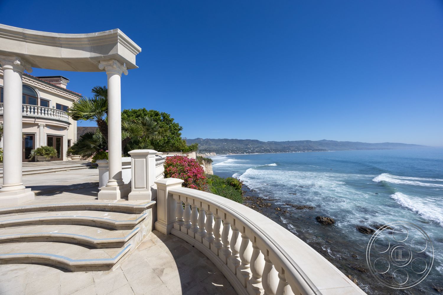 Pacifica Estate - exterior, landscaped garden, large glass windows, arched doorways, plaster columns, Mediterranean architectural style, beachfront view, palm trees landscaping, sunlit outdoor area, luxurious outdoor living space, oceanfront terrace, expansive ocean view, marble patio flooring, balustrade railings, curved stair steps, coastal property, stone-clad foundation, stained wood door