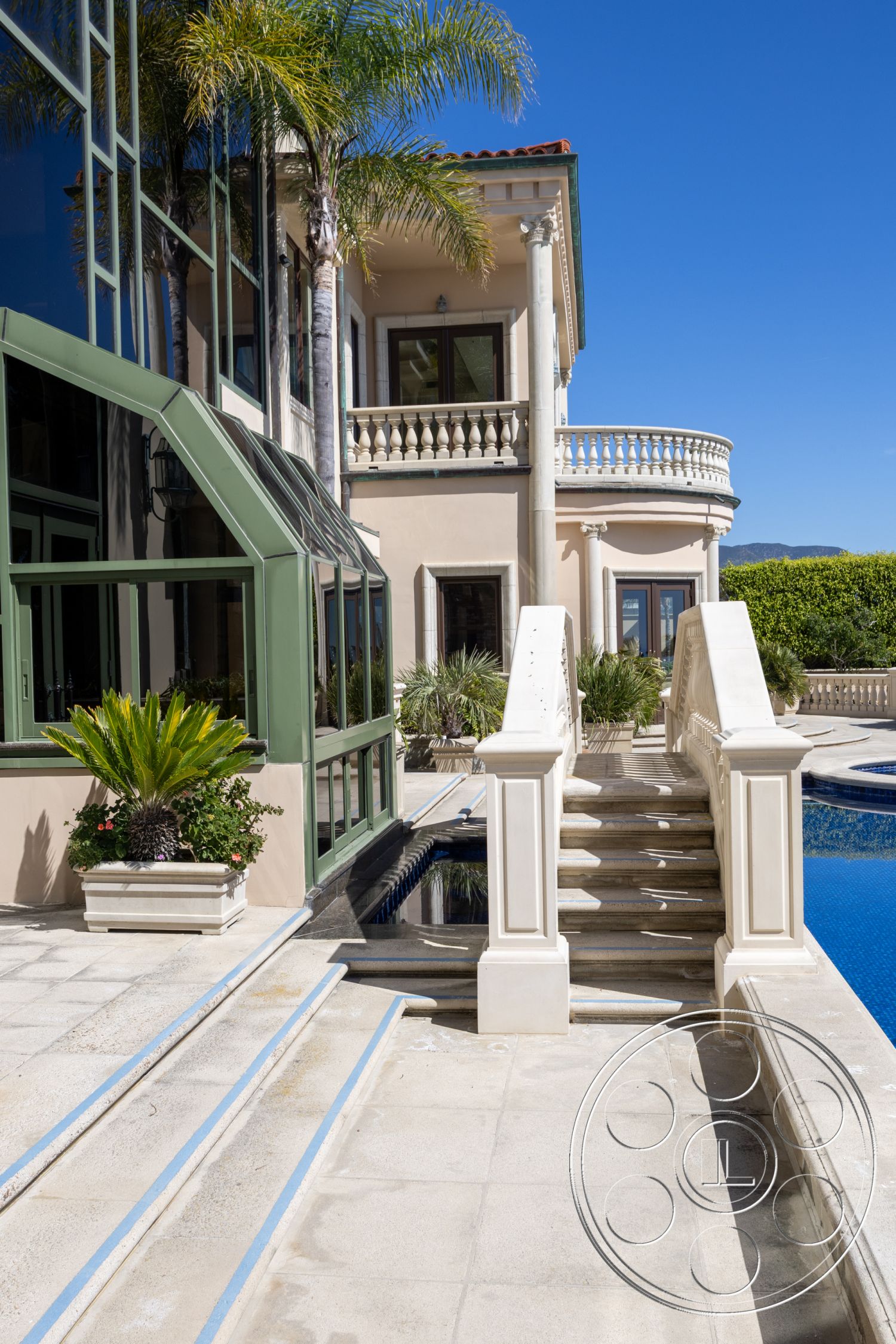 Pacifica Estate - exterior, rectangular swimming pool, stone retaining wall, large sliding glass doors, smooth concrete patio floor, potted plants on patio, marble pool deck, detailed molding around windows, painted concrete steps with decorative railings, exposed column details, glass enclosure for outdoor living area, lush tropical landscaping with palm trees, balustrade detailing on second floor balcony, blue tile waterline, bright sunlight with clear blue sky, formal outdoor setting, municipal luxury villa area, white exterior stucco finishes