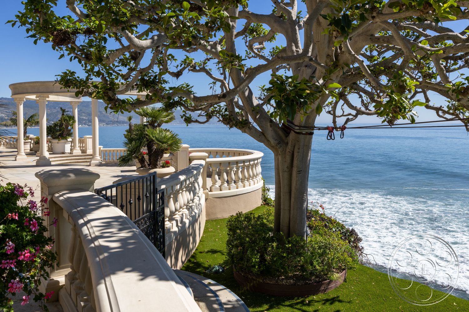 Pacifica Estate - exterior, curved balcony railing, limestone patio flooring, columned terrace with ocean view, planted tropical landscaping, scenic coastal setting, expansive oceanfront deck, exotic wood deck furniture, lush garden area with palm trees, concrete pathway with decorative railing, floral accents along walkway, round gazebo with pillar structure, sunny seaside atmosphere, mature shade tree providing natural canopy, artfully arranged flowering plants, serene blue ocean backdrop, harmonious blend of nature and architecture