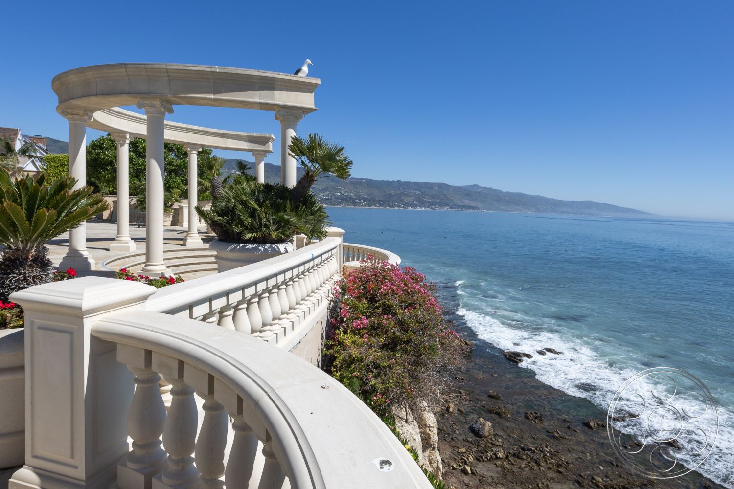 Pacifica Estate - tropical palm trees, outdoor entertainment space, lush greenery surroundings, stone terrace flooring, coastal oceanfront view, clear blue sky backdrop, exterior balcony railing, textured concrete walkway, decorative wrought iron details, ornamental flower beds, sunny seaside setting, planted tropical landscaping, pillared pergola structure, curved balustrade, overlook seating area, classic Mediterranean architectural style, circular column support, smooth finish marble wall, expansive ocean view terrace, coastal cliffside location