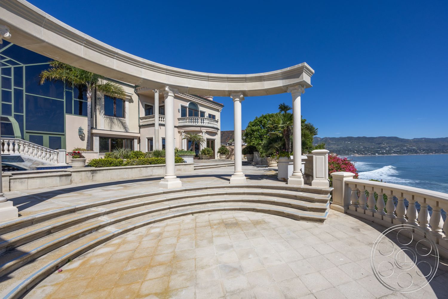 Pacifica Estate - exterior, landscaped garden, floor-to-ceiling windows, arched doorways, sunny coastal setting, coastal view, marble columns, modern mansion, modern Mediterranean style, stone patio flooring, curved patio, palm trees in garden, balustrade railing, oceanfront deck, luxury outdoor entertaining area, smooth concrete terrace, sweeping ocean views, expansive patio space