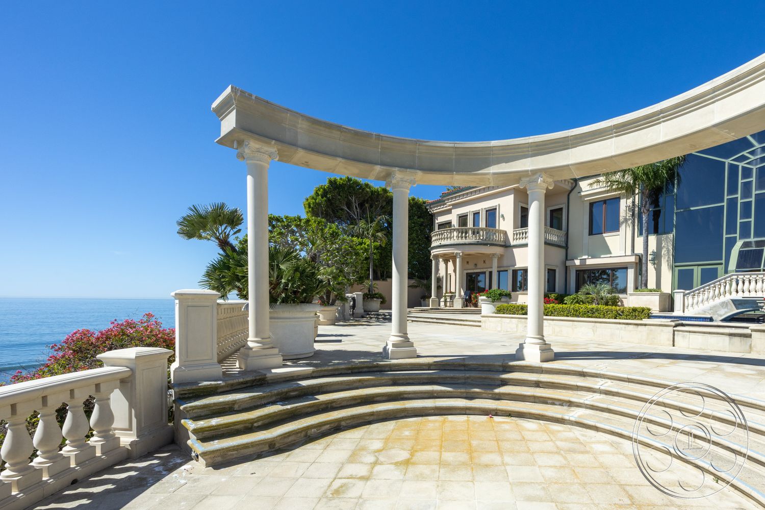 Pacifica Estate - outdoor living space, open-air terrace, large floor-to-ceiling windows, stone patio flooring, exterior terrace with ocean view, modern Mediterranean architectural style, rectangular entryway, landscaped garden with palm trees, curved decorative colonnade, white plastered exterior walls, lattice balcony railing, ornamental stone columns, flowering shrubs and bushes, circular steps leading down, sweeping coastal views