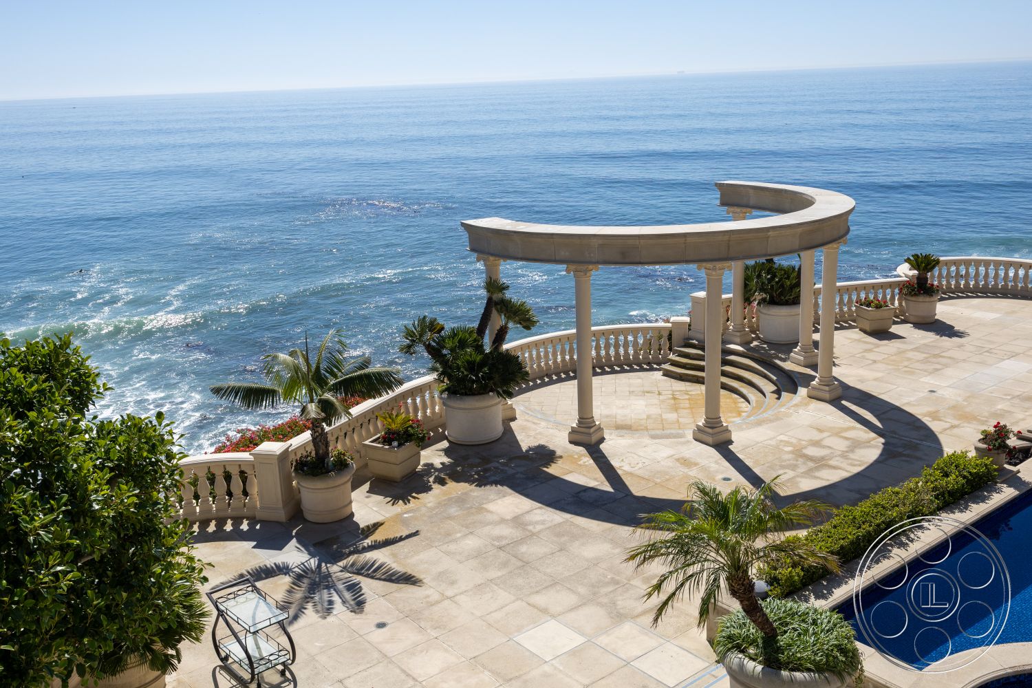 Pacifica Estate - natural light, open-air design, tropical landscaping, outdoor lounging area, blue water pool, stone patio flooring, oceanfront view, exterior patio flooring, mediterranean architectural style, sun-drenched terrace, curved colonnade, circular colonnade structure, marble poolside coping, planted garden pots, smooth stone pillars, decorative balustrade, serene coastal setting, soft shadows from palm trees, staircase with multiple steps, expansive ocean vista