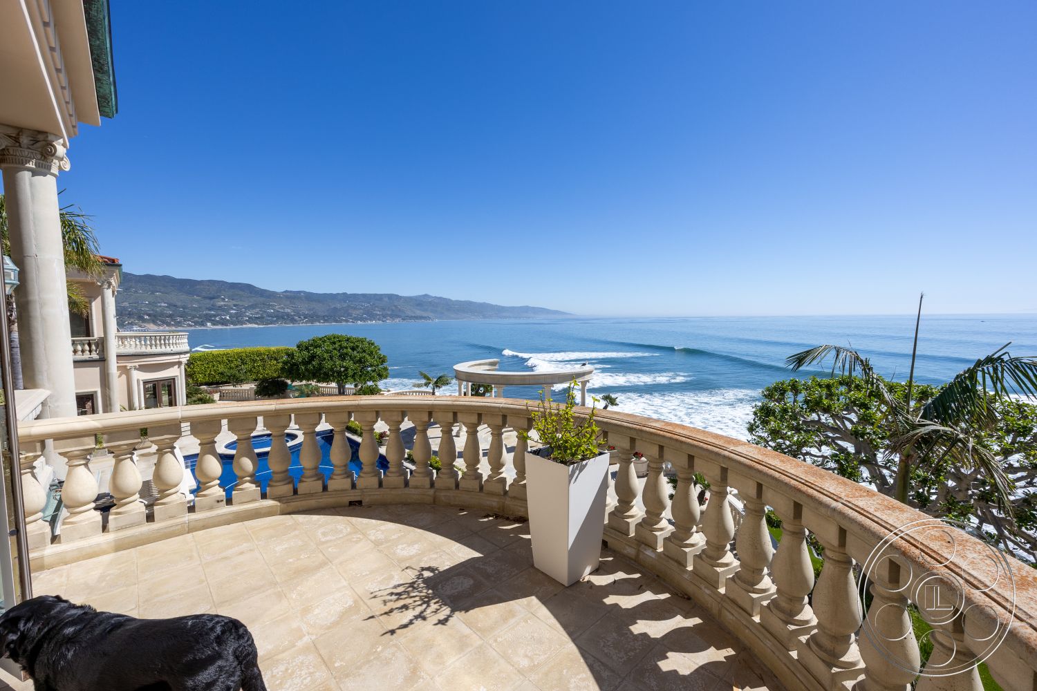Pacifica Estate - clear blue sky, Mediterranean architectural style, landscaped garden area, sandy beach access, sunny hillside location, palm trees in foreground, exterior balcony with stone tile flooring, curved balcony railing with balustrade details, oceanfront view with coastal landscape, luxury coastal home architecture, columned support structures, infinity pool in background, terracotta planter accents