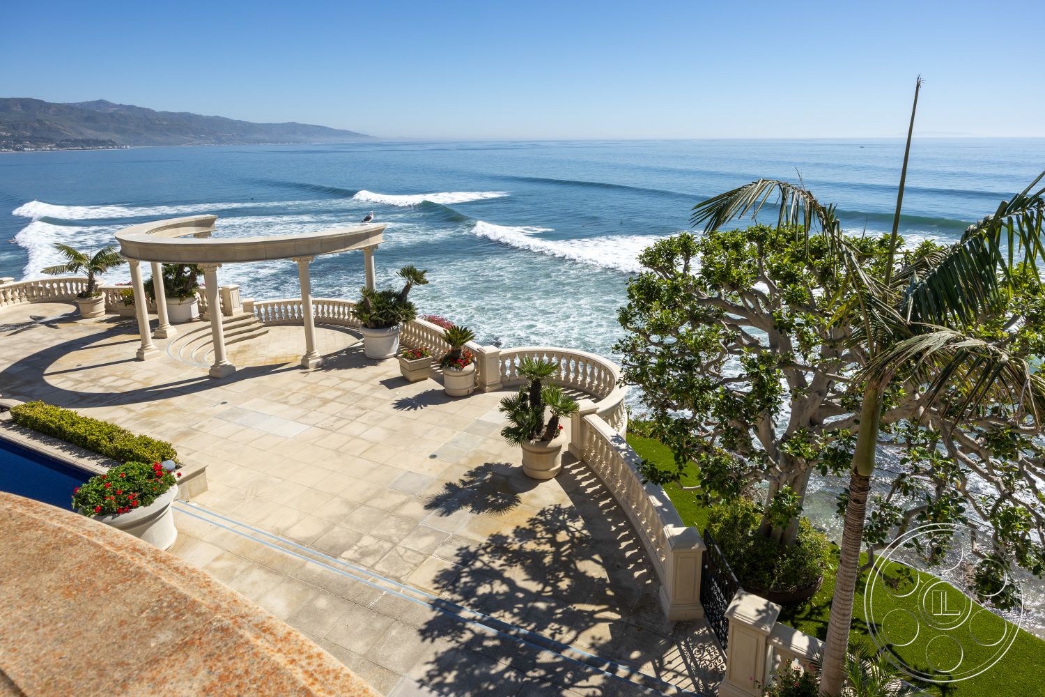 Pacifica Estate - smooth concrete surface, outdoor living space, arched entrance, mediterranean architecture, stone patio flooring, palm tree landscaping, exterior patio flooring, coastal oceanfront view, infinity pool edge, curved landscaping hardscape, white classical columns, planter boxes with flowers, overlook terrace, lush grass landscaping, scenic beachside setting