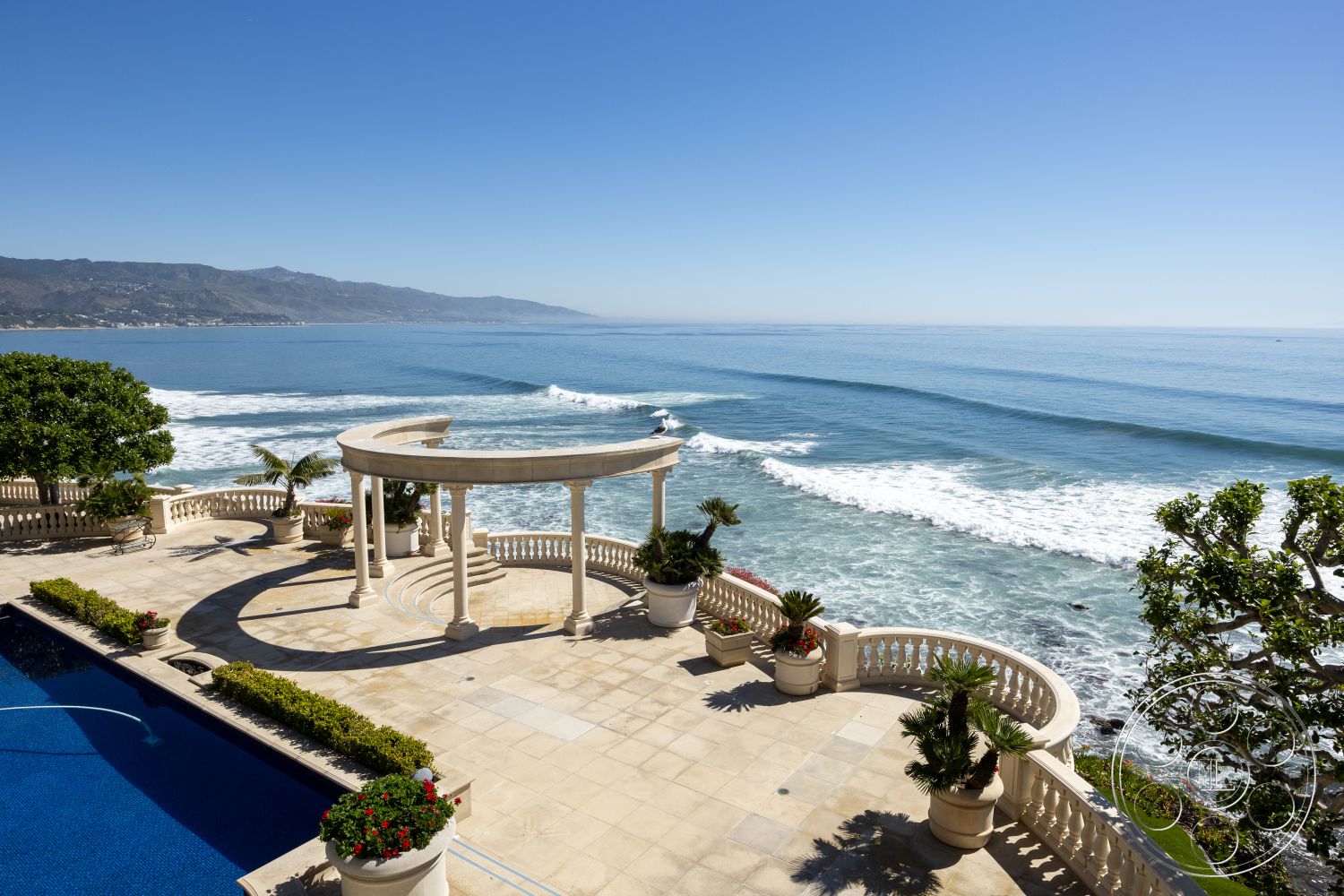 Pacifica Estate - rectangular swimming pool, natural stone walls, outdoor lounging area, coastal setting, modern coastal architecture, landscaped garden beds, stone patio flooring, oceanfront view, exterior pool area, marble pool deck, curved colonnade structure, white balustrade railing, tropical landscape with palm trees, rounded garden planters, sunny outdoor terrace